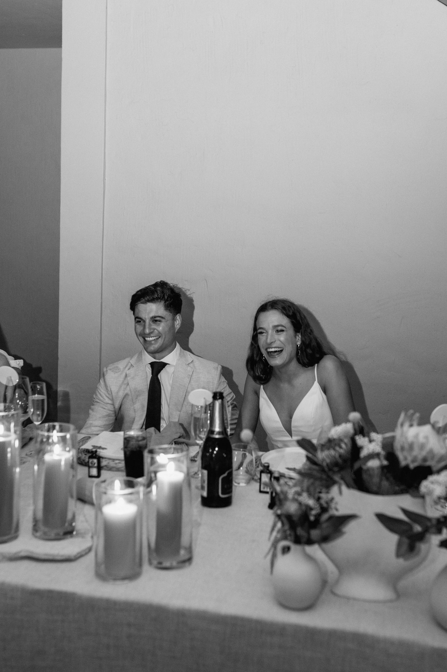 Black and white photo of wedding guests laughing at head table with candles and drinks.