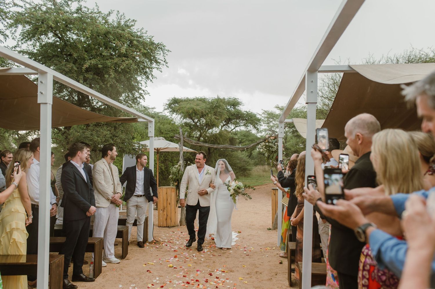 Wedding guests gather under brown canopies along a rustic outdoor path during a ceremony.