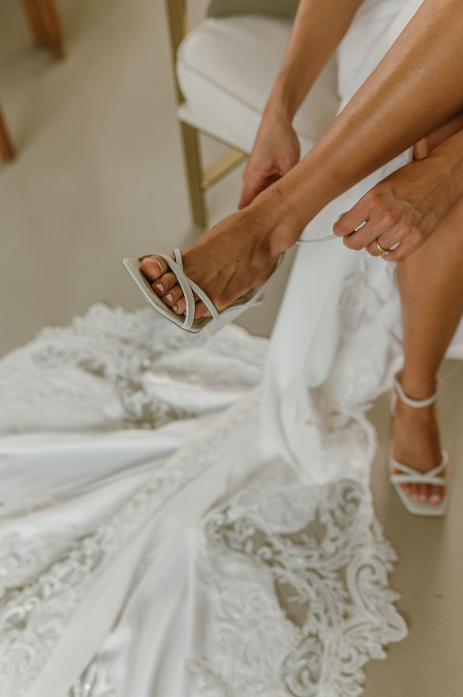 Close-up of white wedding shoes being fastened with lace dress details visible.