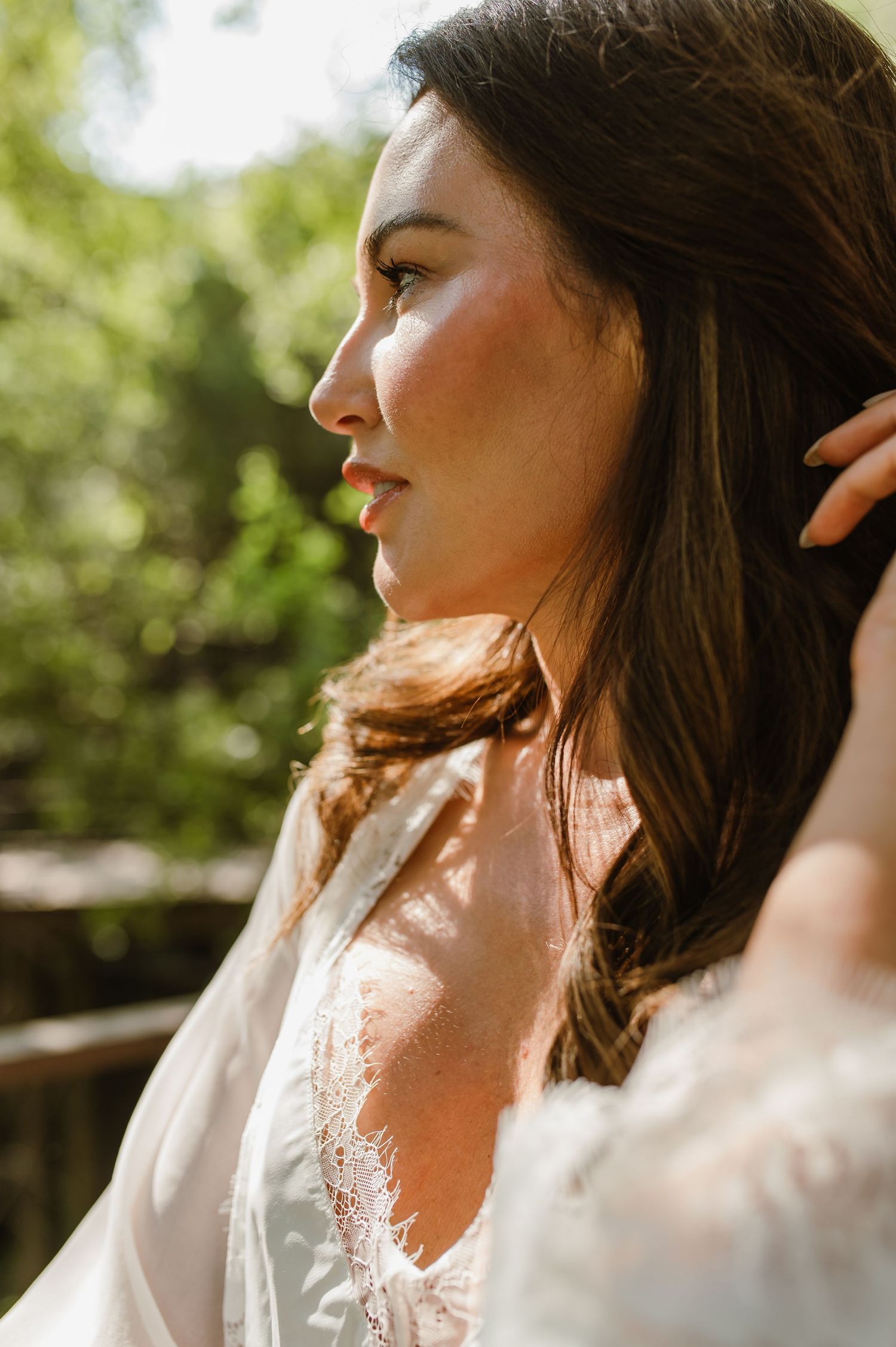 Close-up profile of brunette bride in white lace robe with sunlight filtering through trees outdoors.