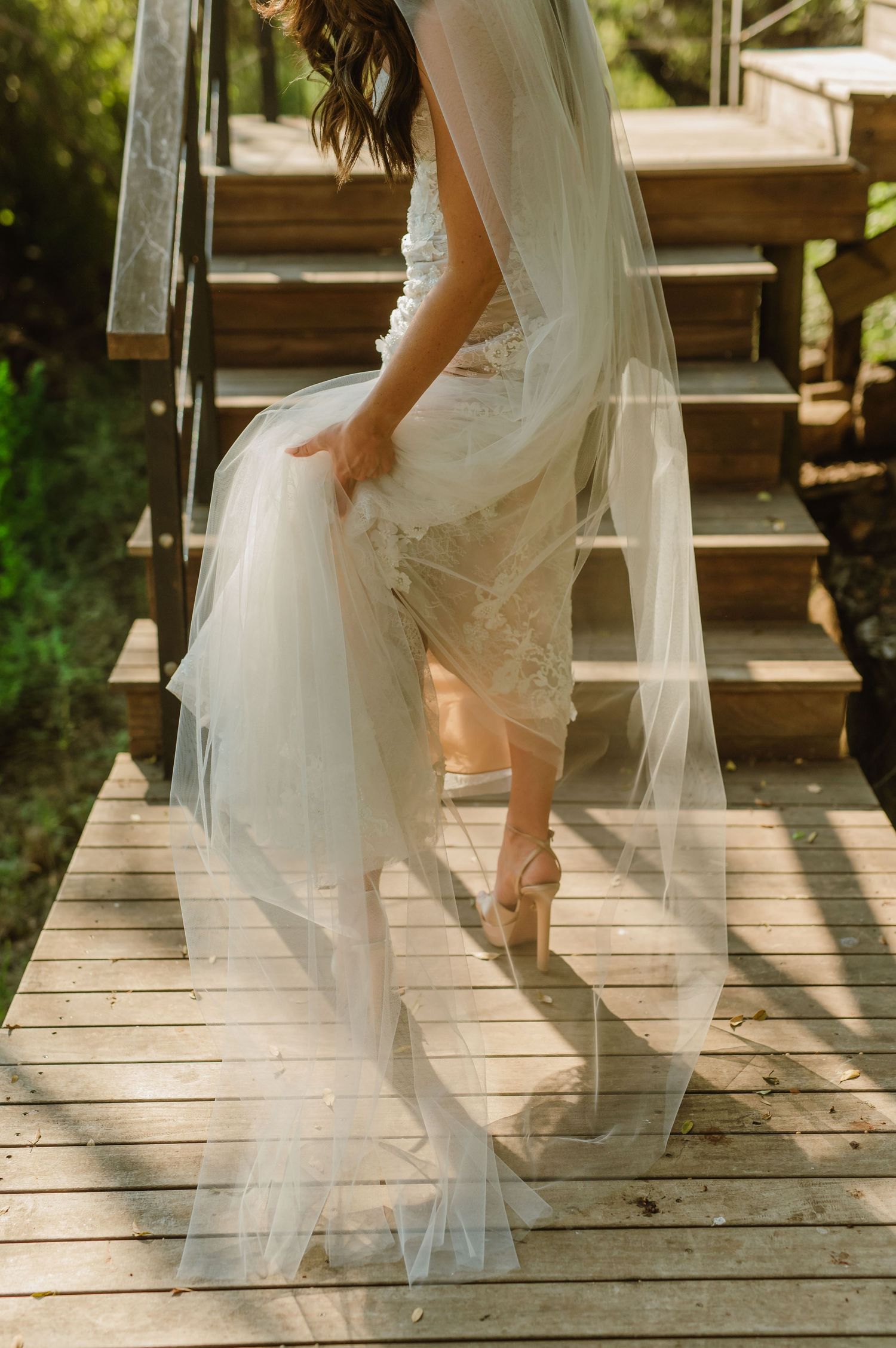 Bride in lace wedding dress descending wooden stairs holding veil with strappy heeled sandals.