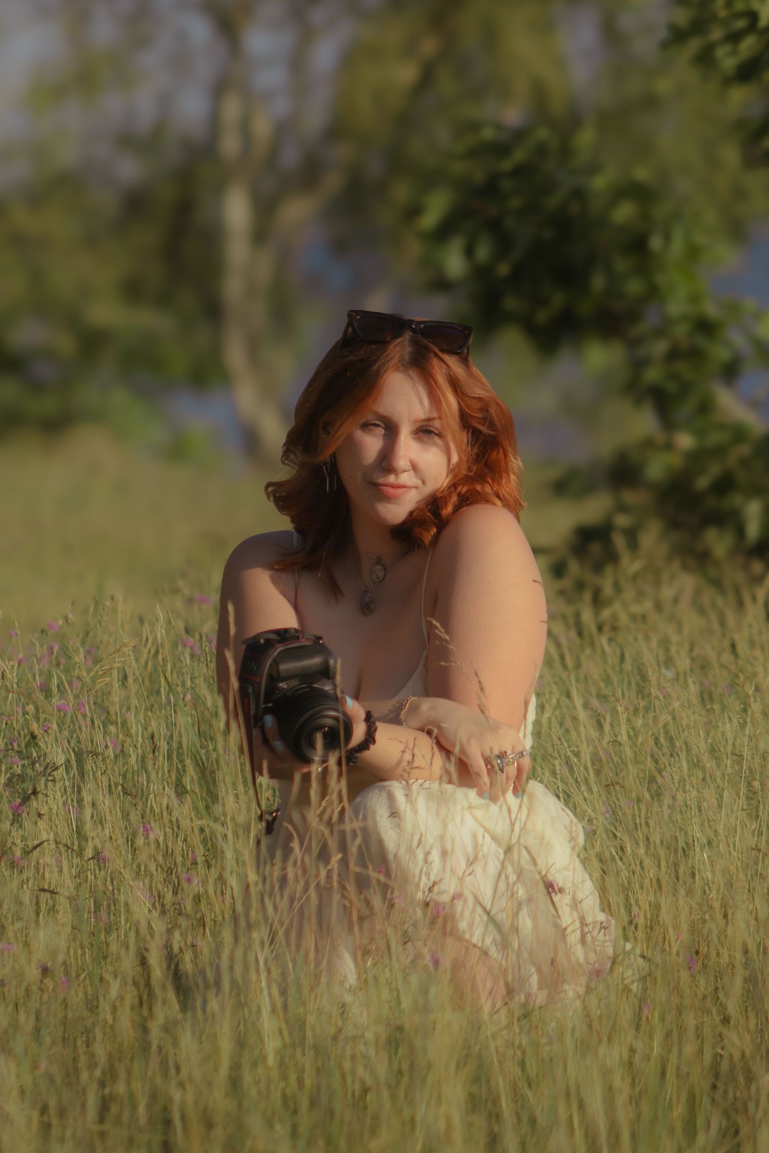Red-haired woman sitting in green meadow holding vintage camera, wearing white dress with sunglasses on head.