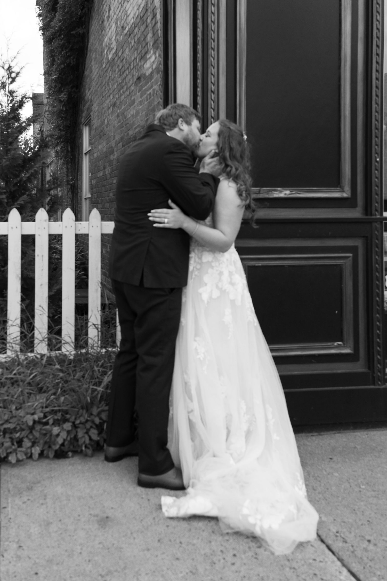 A wedding couple shares an intimate moment near a white picket fence and brick building.