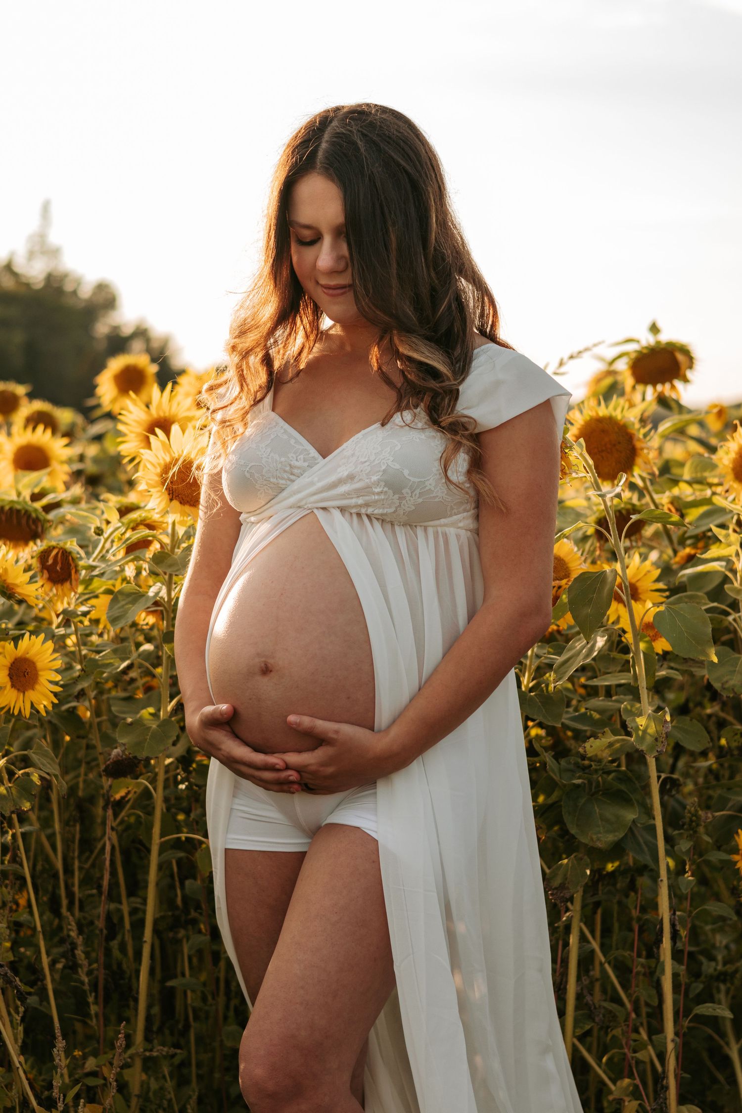Artistic maternity portraits in white dress among blooming sunflowers at sunset.