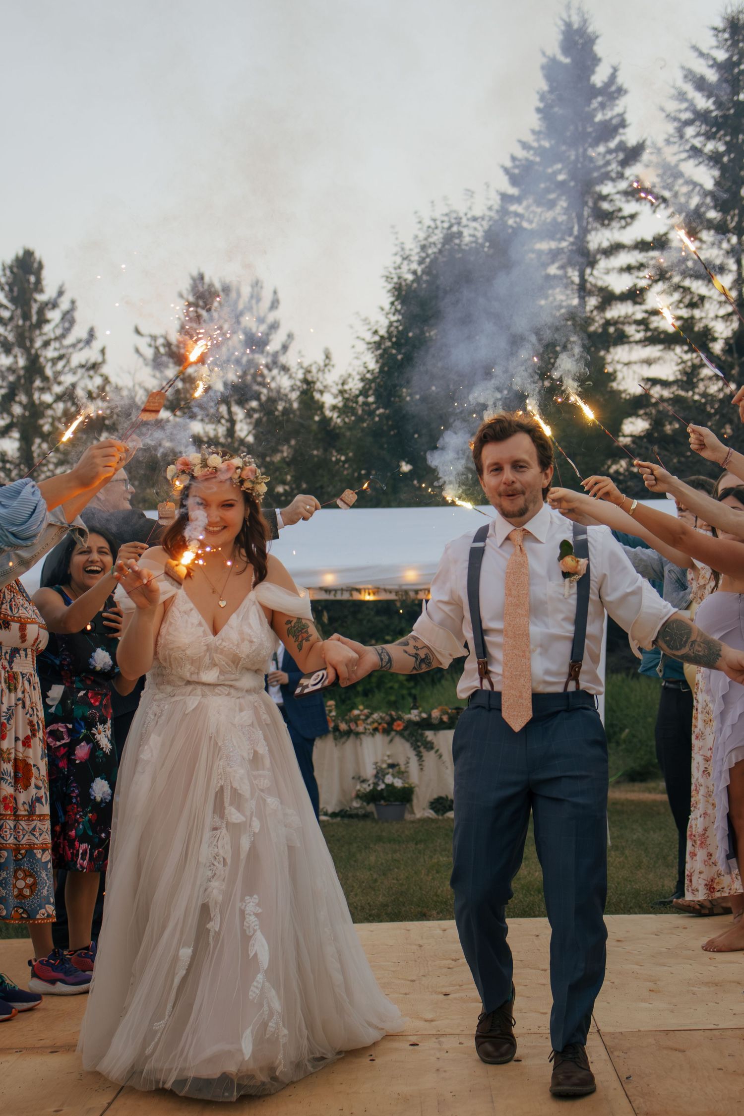 Bride and groom walk through sparkler exit at outdoor wedding ceremony at dusk with guests cheering.
