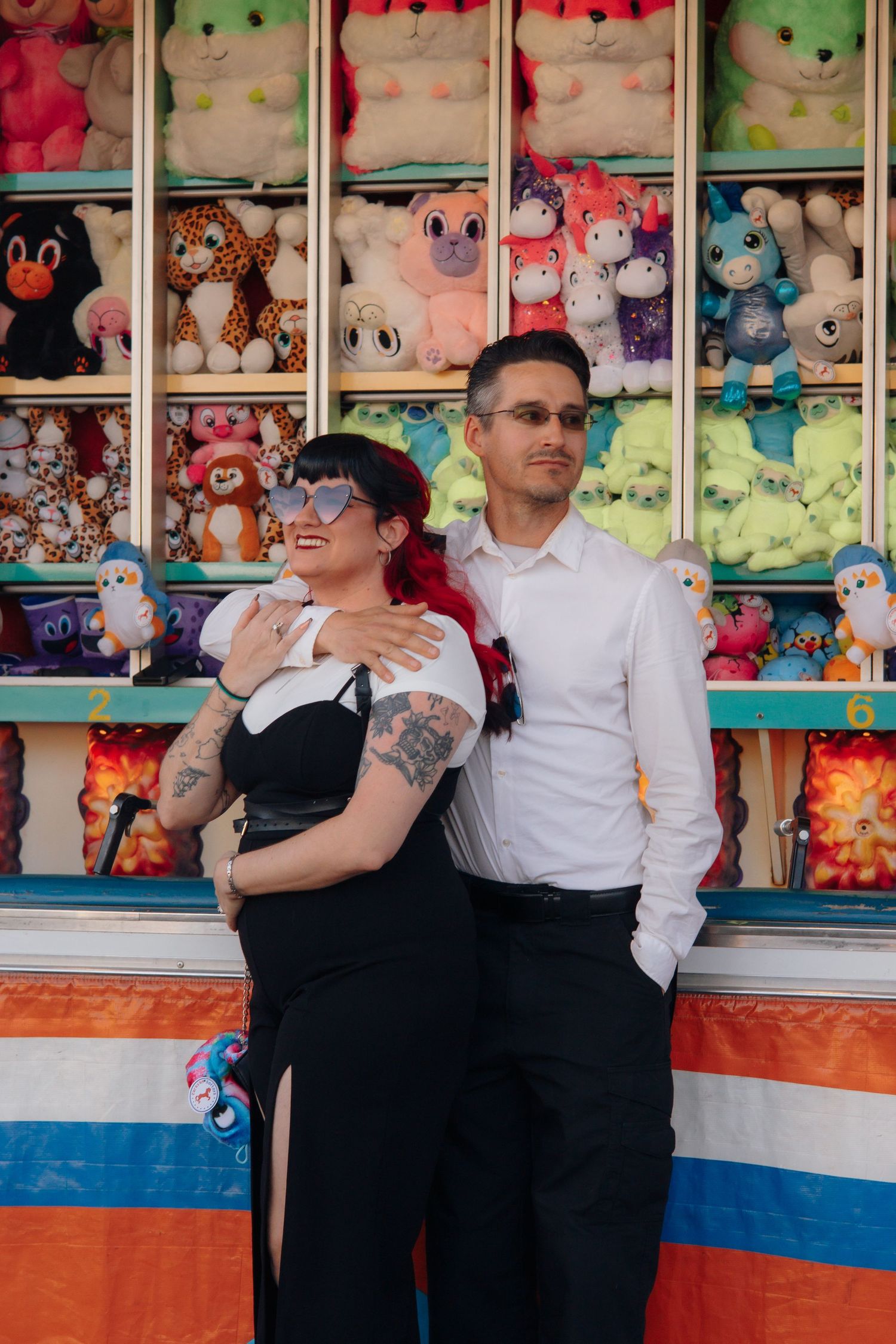 Couple posing in front of colorful carnival prize booth filled with stuffed animals at a fair.