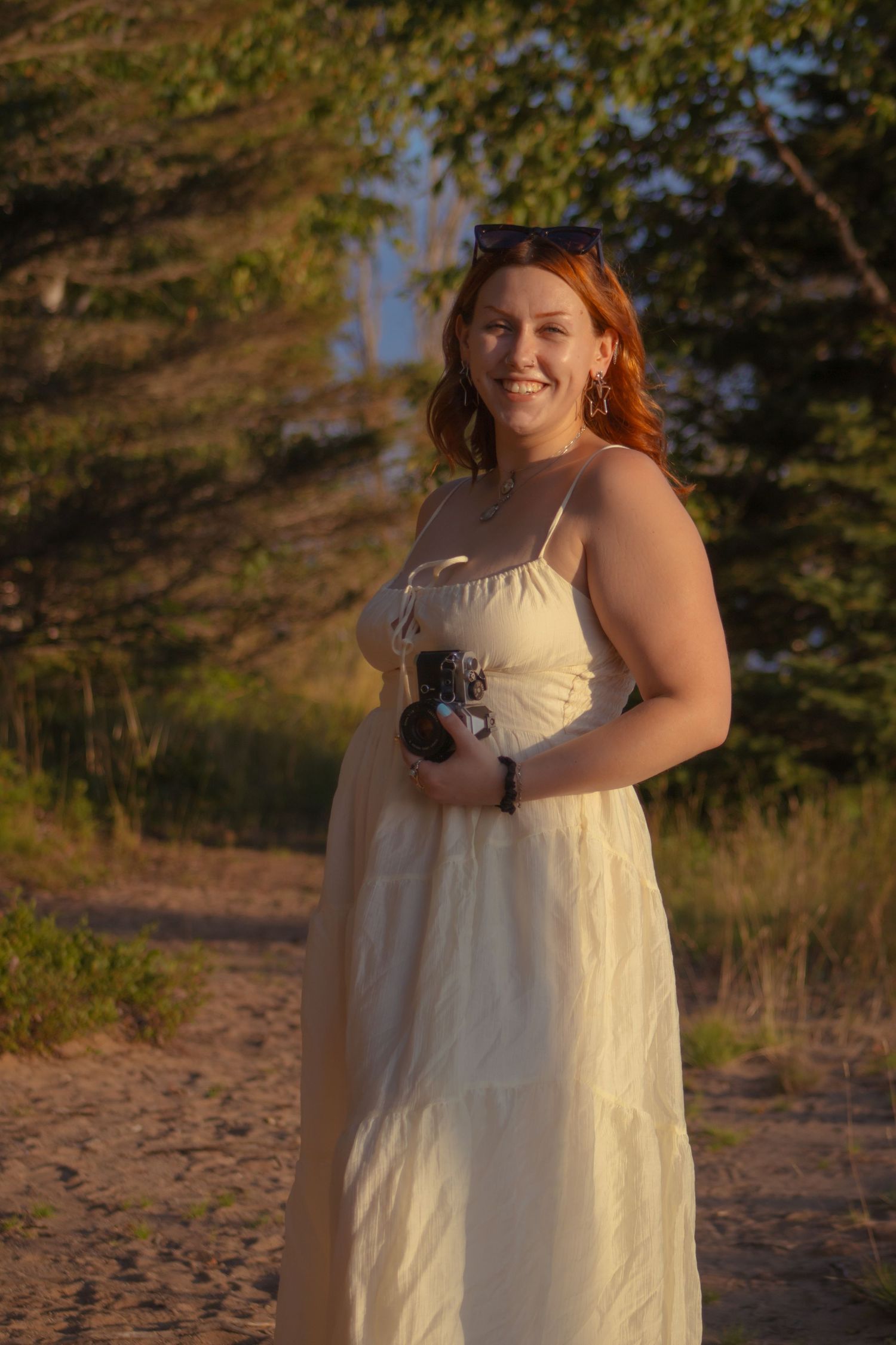 Smiling redhead woman in white sundress holding camera outdoors during golden hour on sandy path.