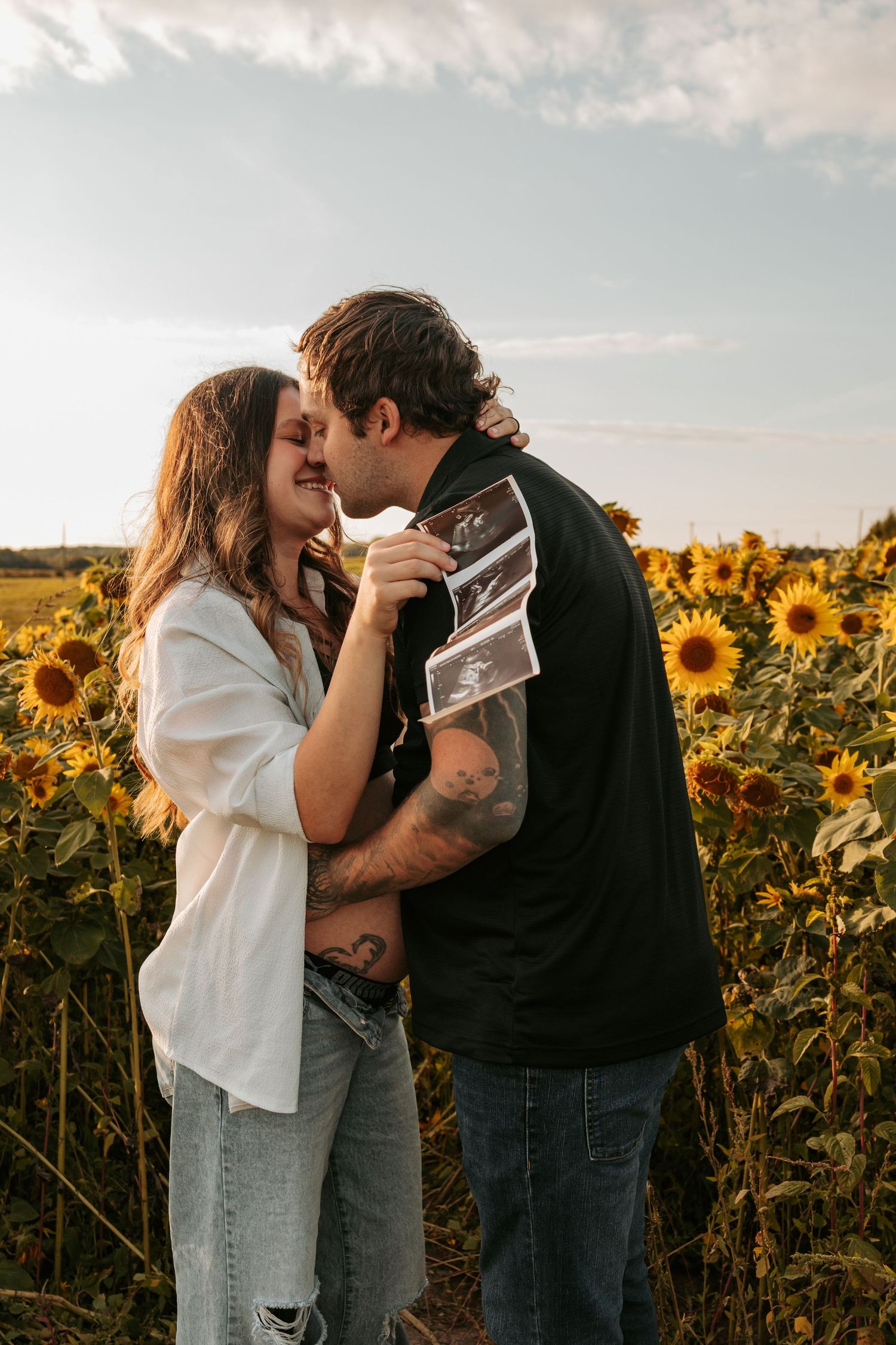 Couple shares tender moment in sunflower field while holding ultrasound photo during sunset.