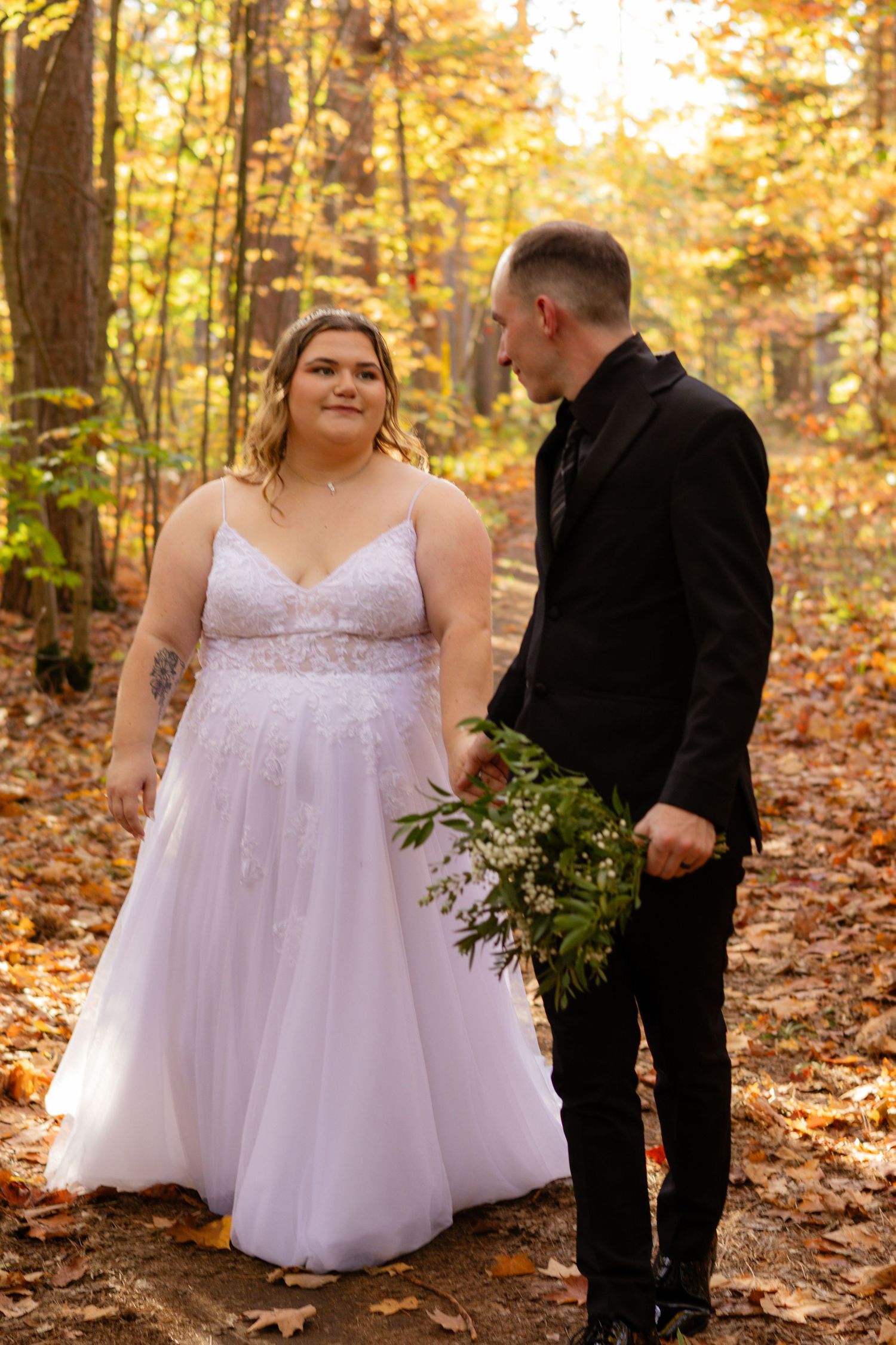 A wedding couple stands together on a leaf-covered forest path during golden hour.