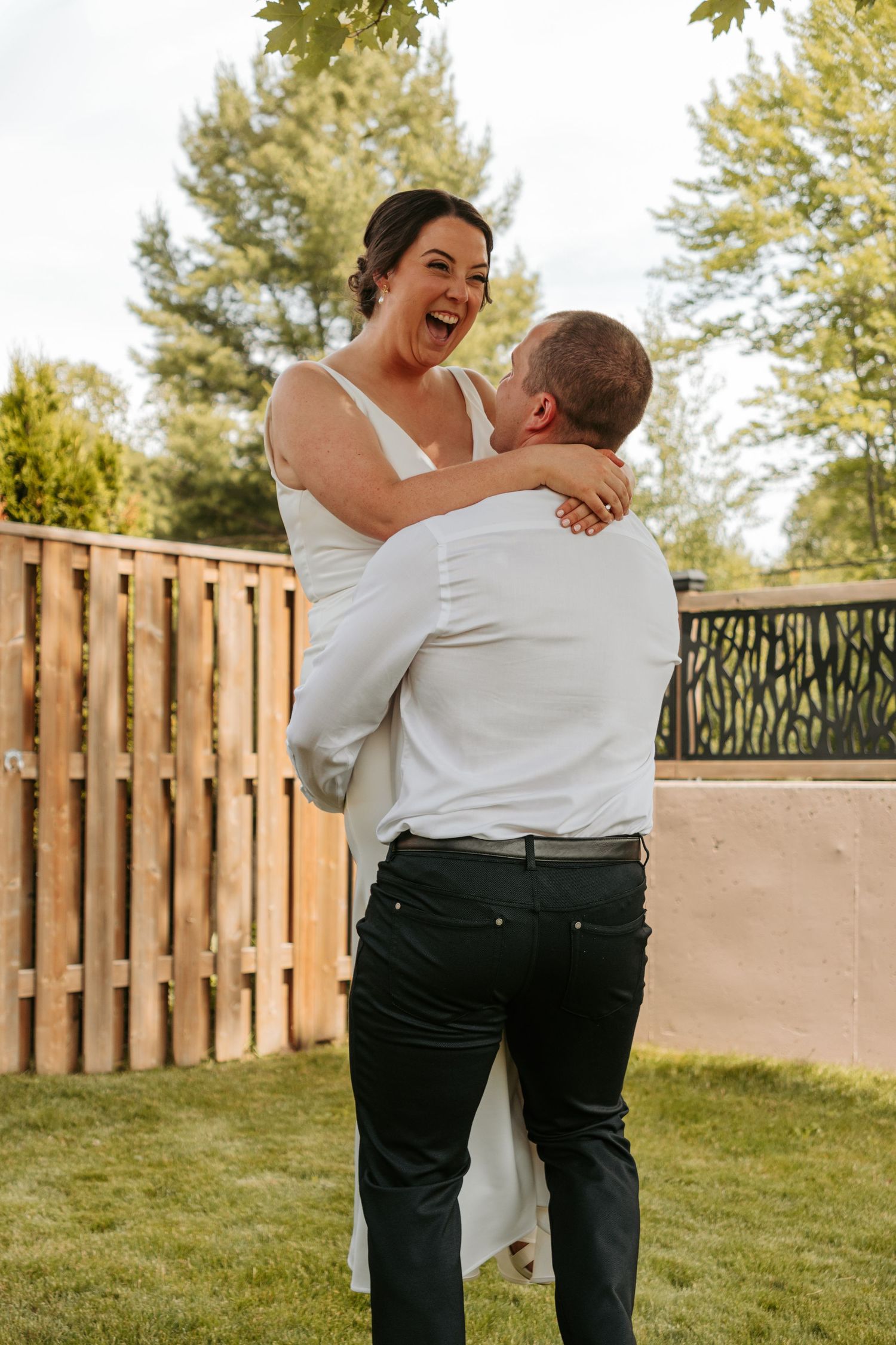 A joyful wedding couple embraces and laughs together on a lawn with wooden fencing in the background.