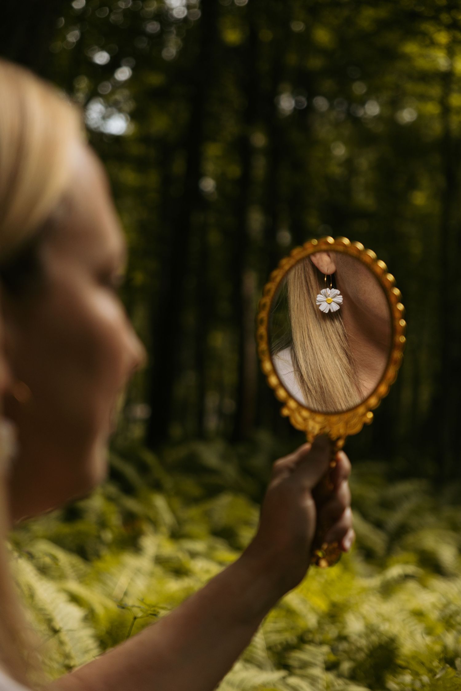 Hand holding vintage mirror reflecting jewelry and blonde hair in forest setting.