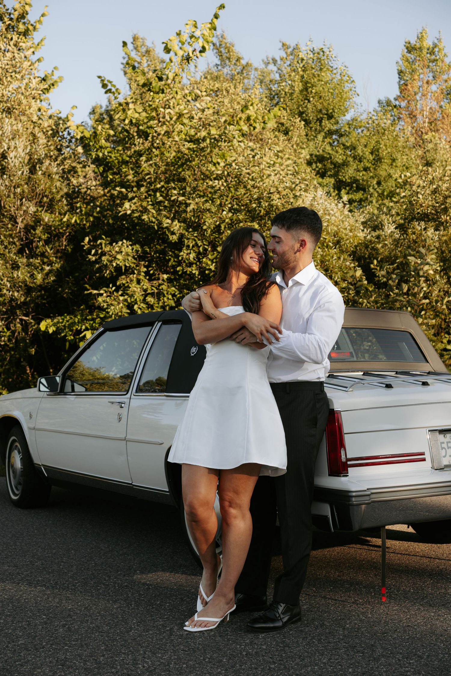 Couple in white and black outfits pose romantically by vintage car.