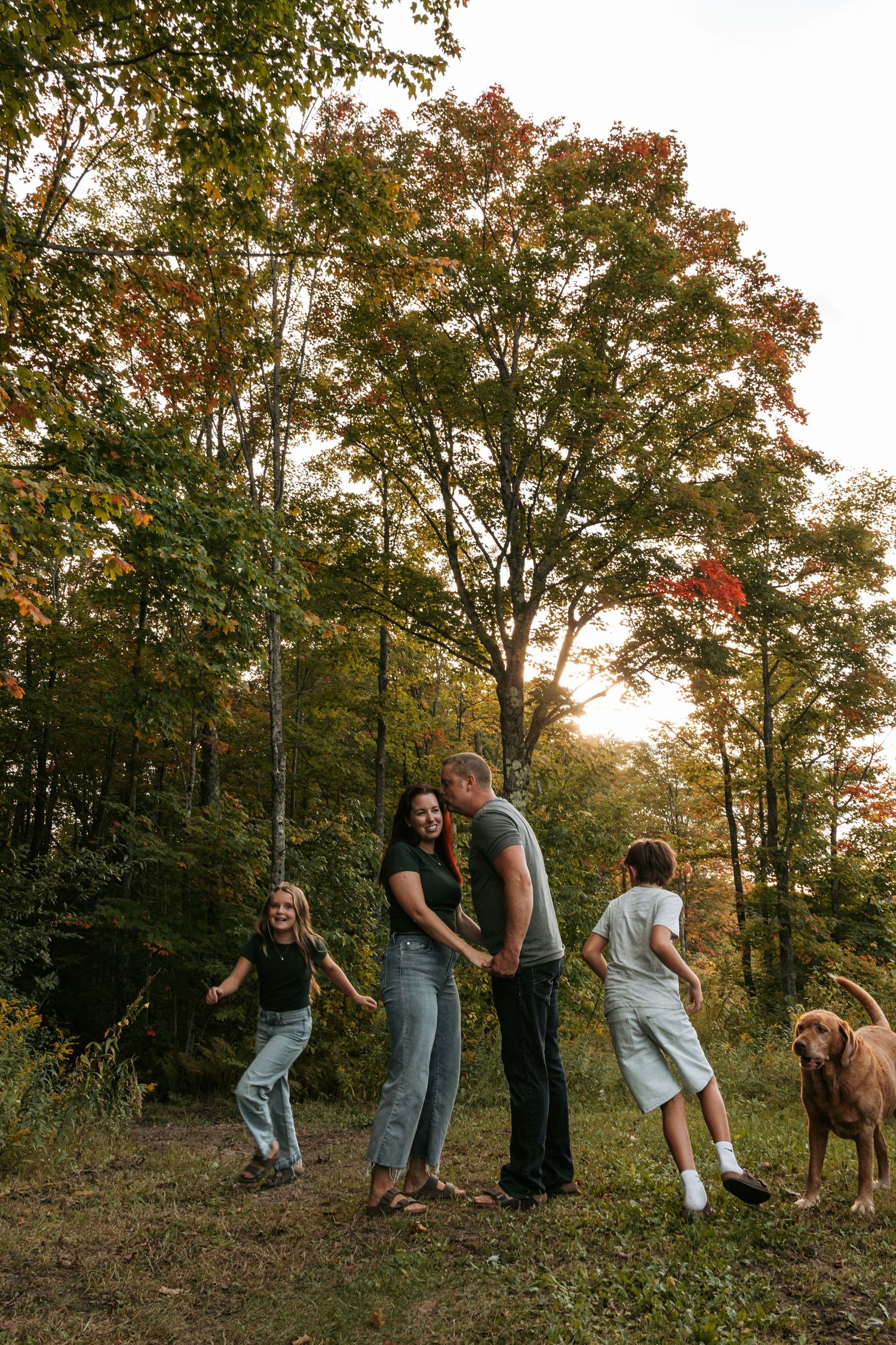 Family and dog enjoying a playful moment together on a wooded hillside during sunset.