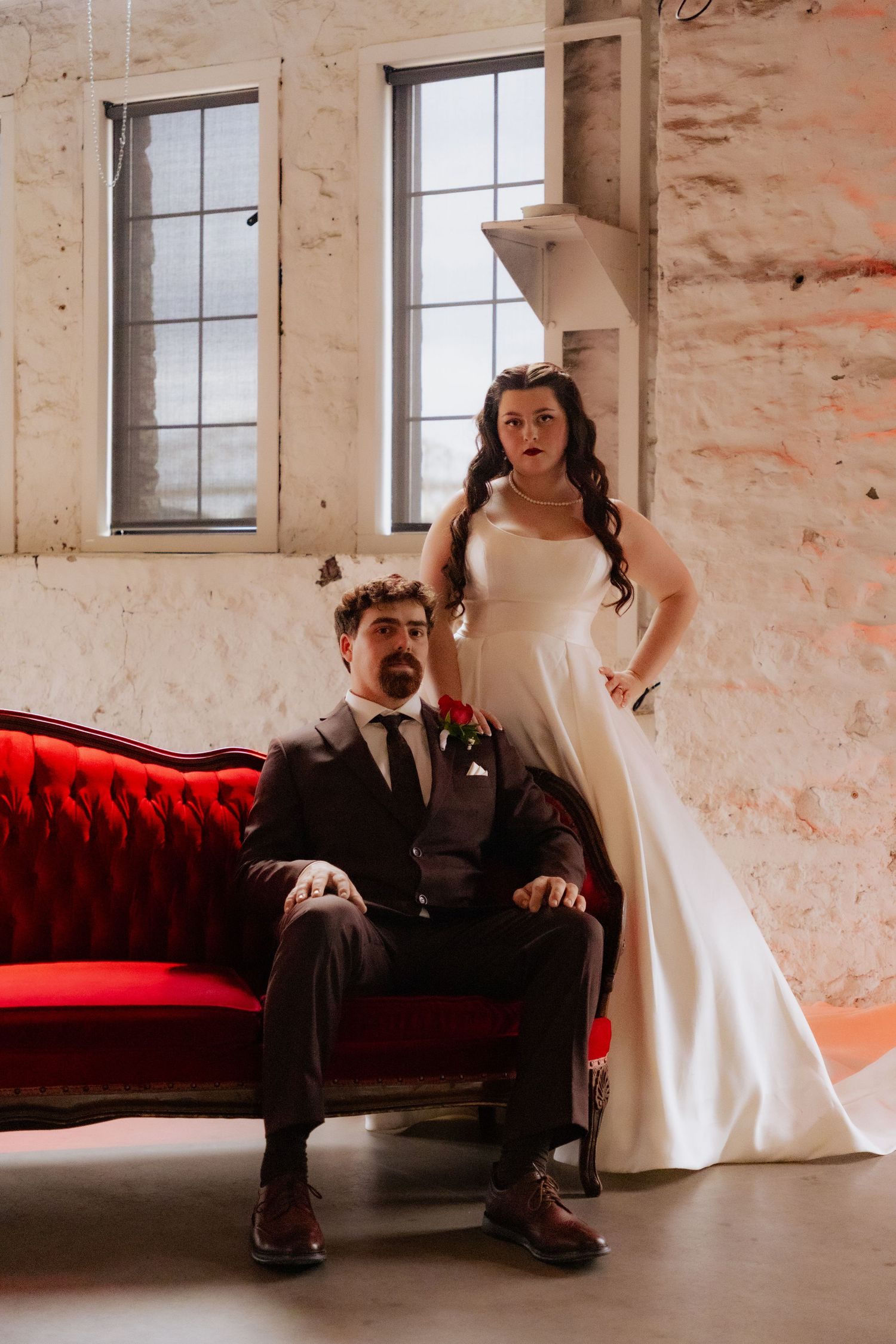 A wedding couple poses on a red velvet couch in an industrial brick venue.