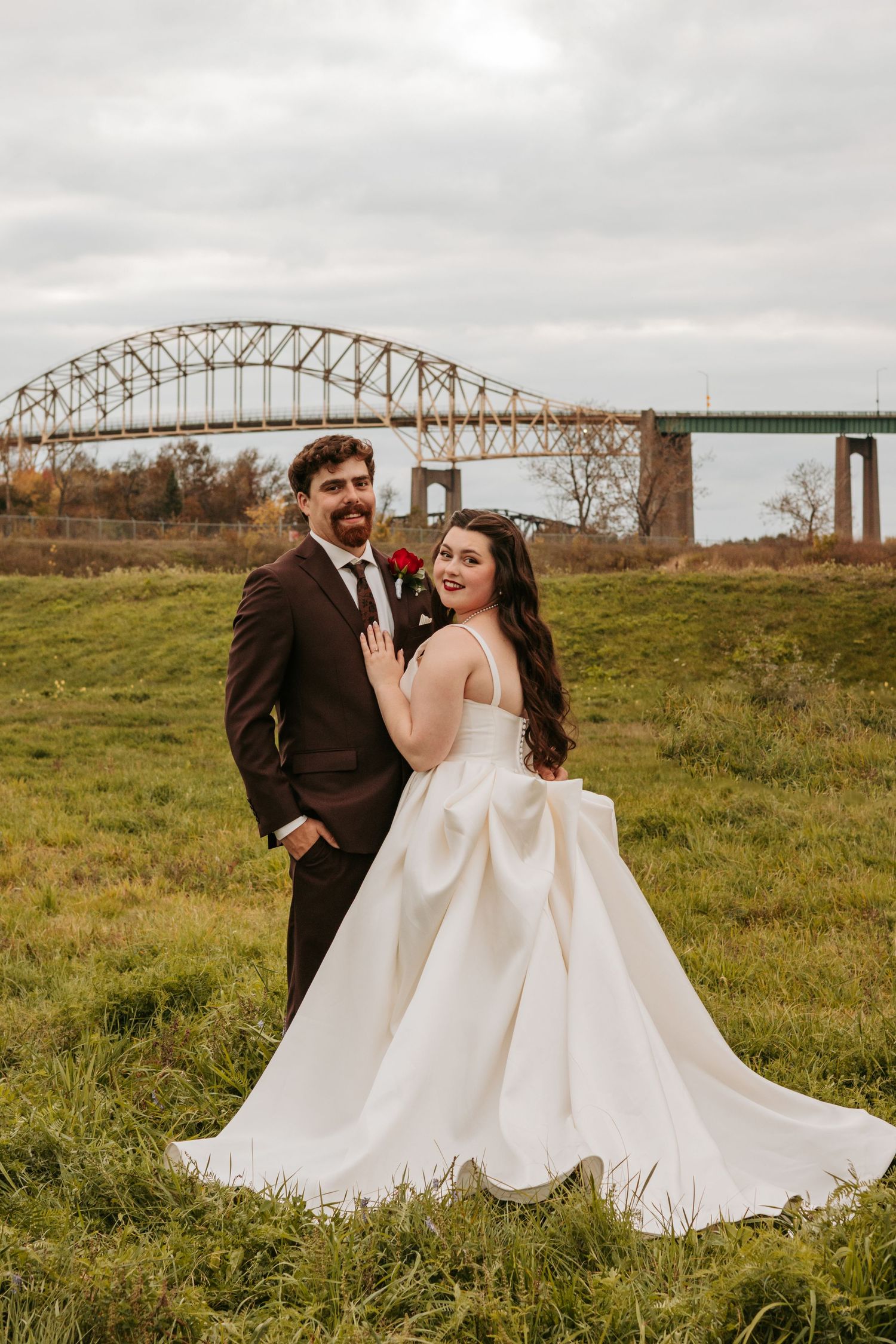 A wedding couple poses in a field with a large bridge structure in the background.
