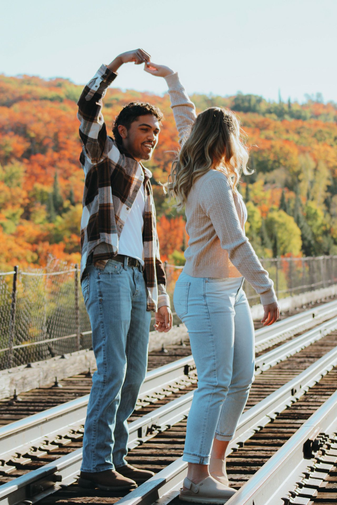 A couple dances playfully on railroad tracks surrounded by vibrant autumn foliage.