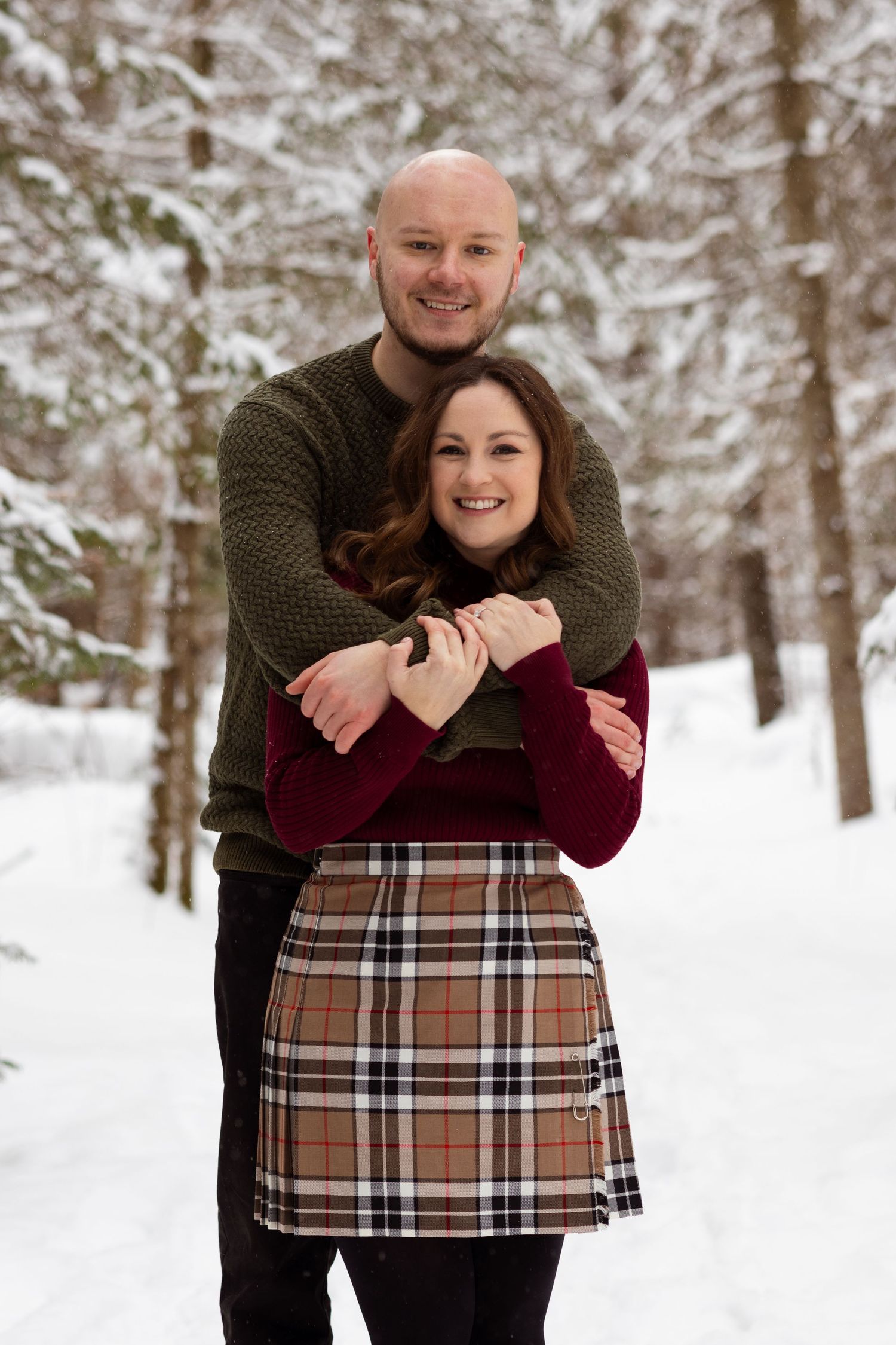 A joyful couple poses in a snowy forest setting, with her wearing a plaid skirt and burgundy sweater.