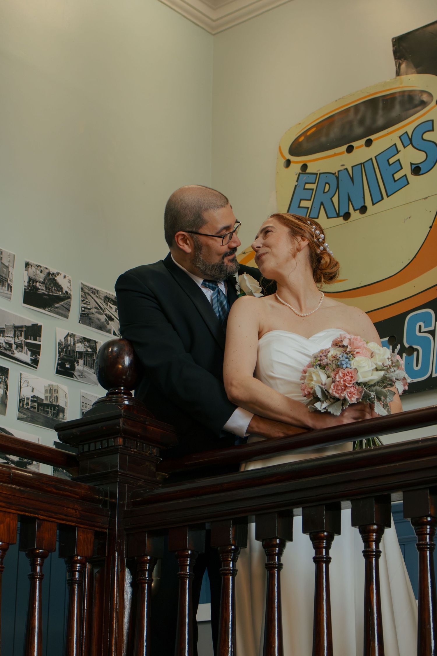 Bride and groom share a romantic moment on a wooden balcony near a vintage Ernie's sign on their wedding day.