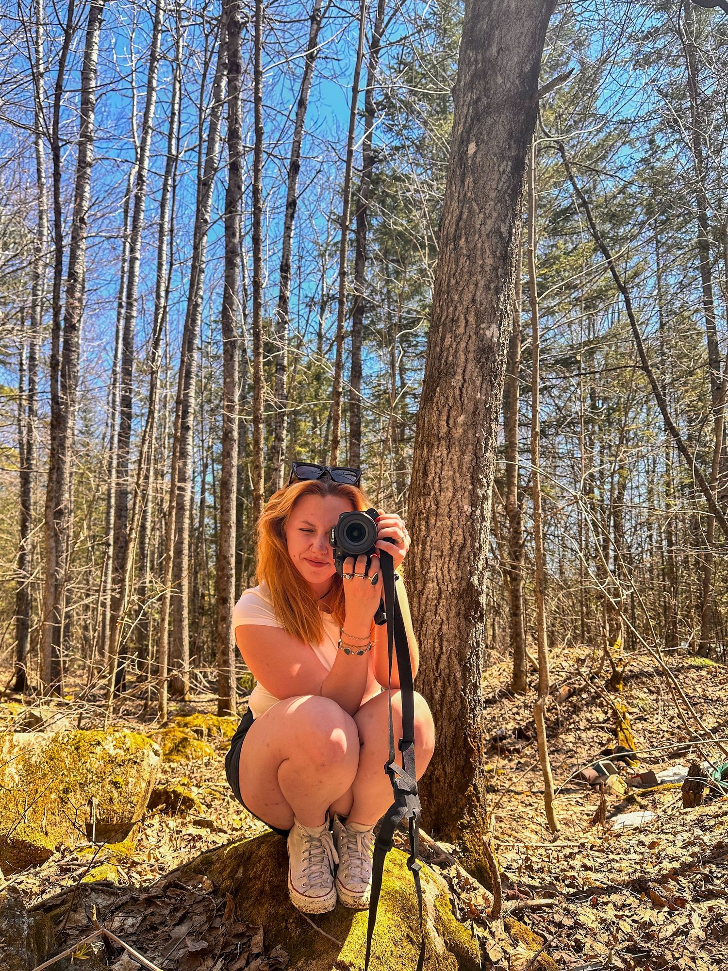 A red-haired woman crouches in a sunlit forest, smiling while holding a camera on a tripod among bare trees.