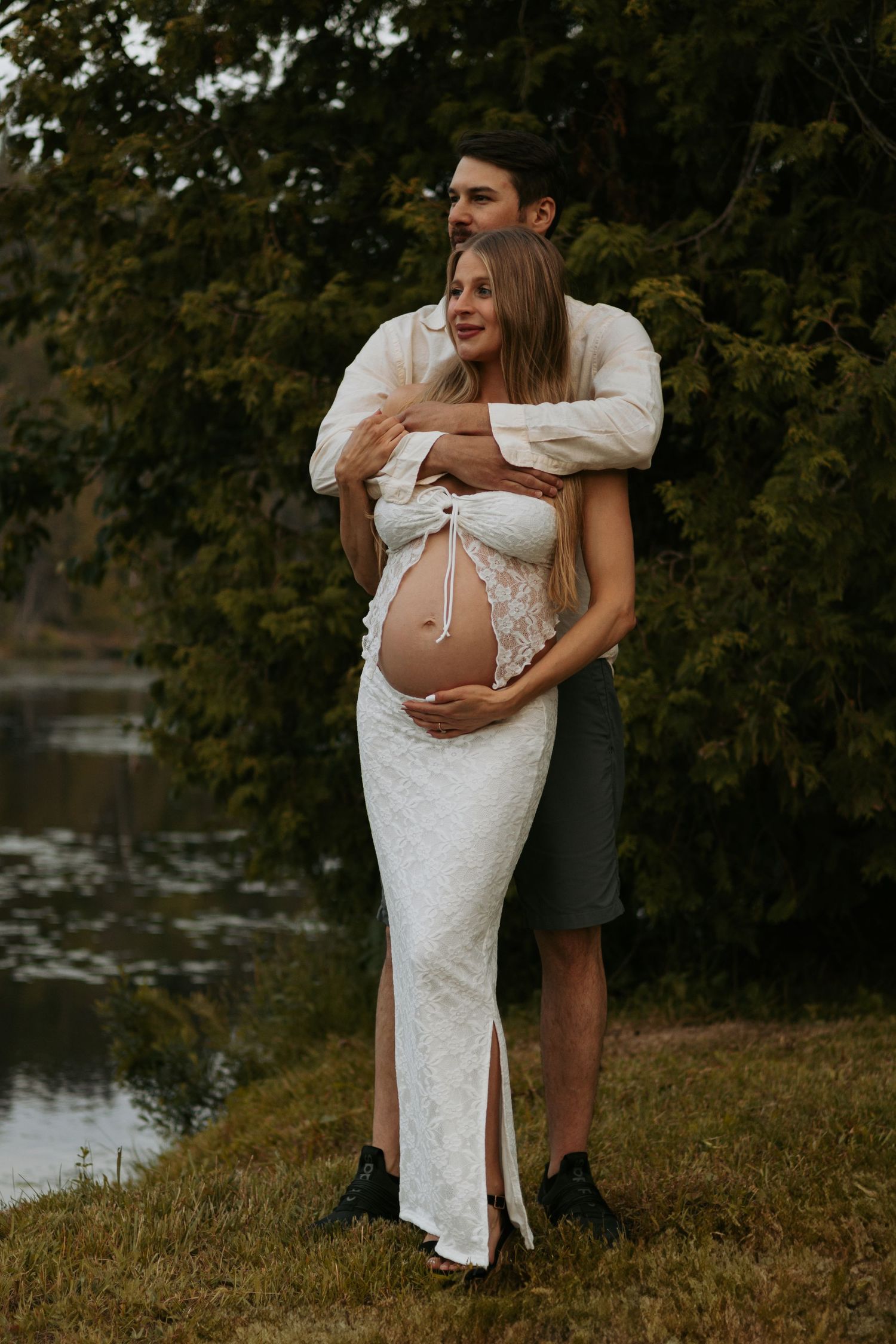 Maternity photos showing couple embracing near lake in white lace dress.