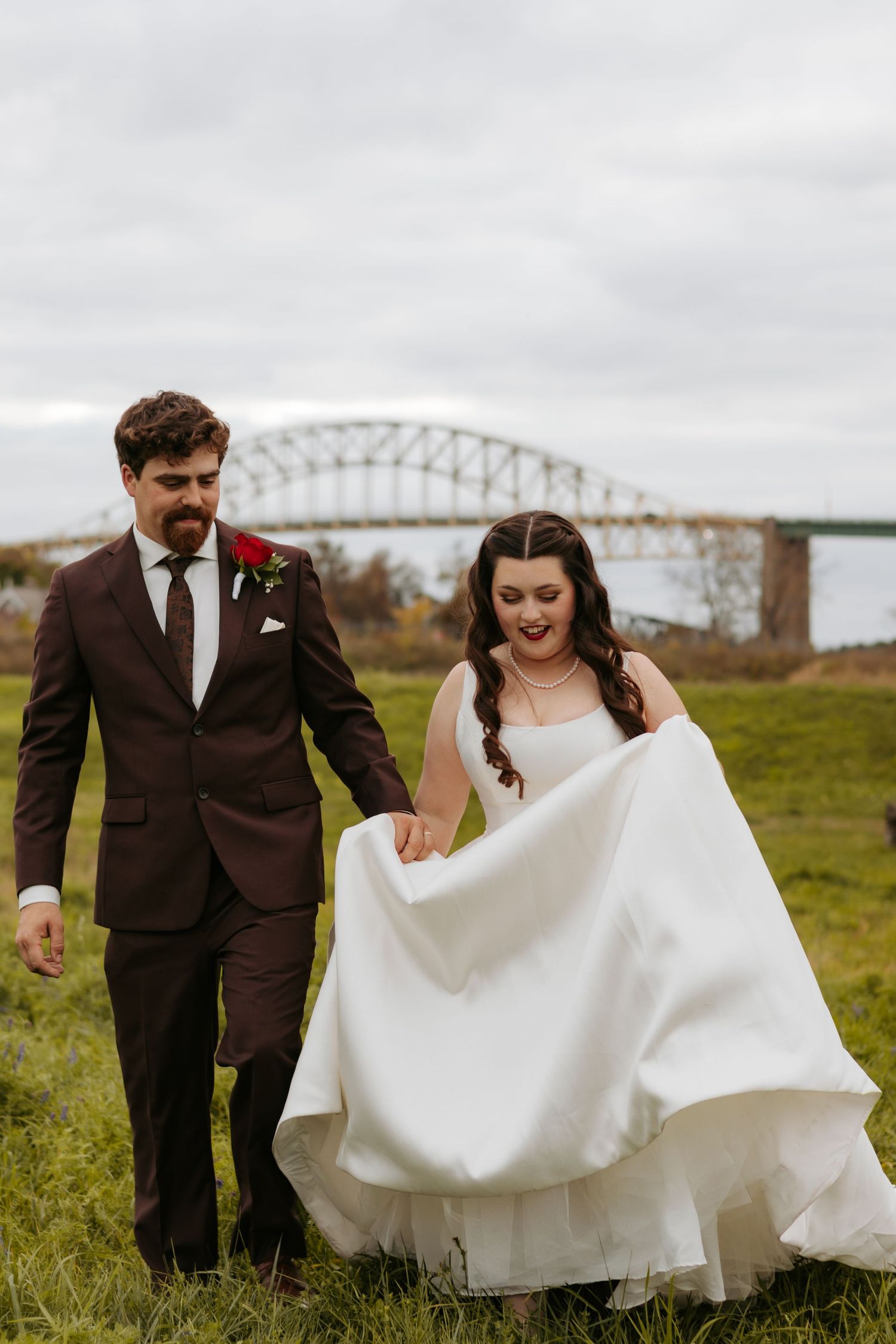 A couple walks through a grassy field with a bridge in the background on their wedding day.