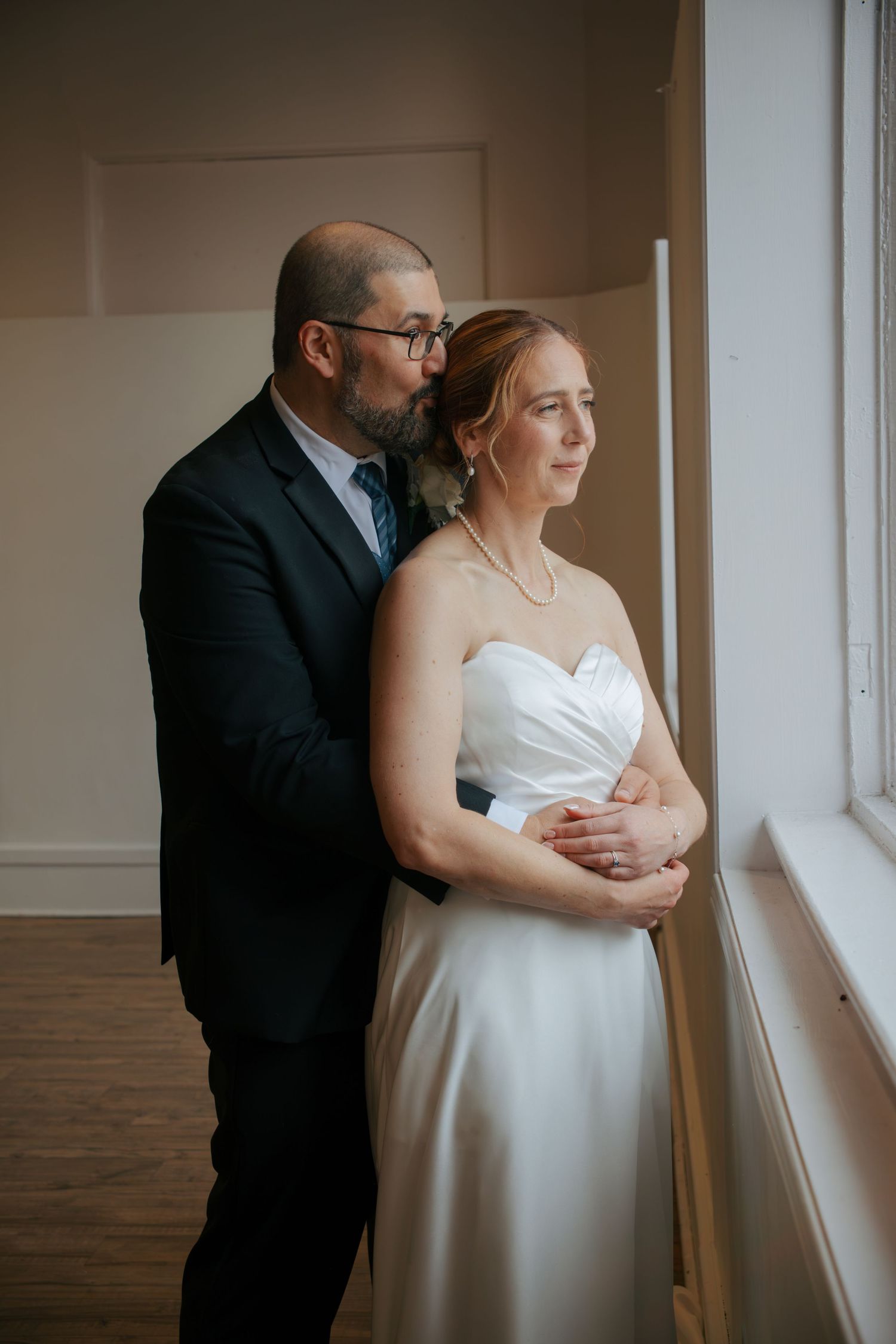 A groom in a dark suit embraces his bride in a white strapless gown as they gaze out a window together.