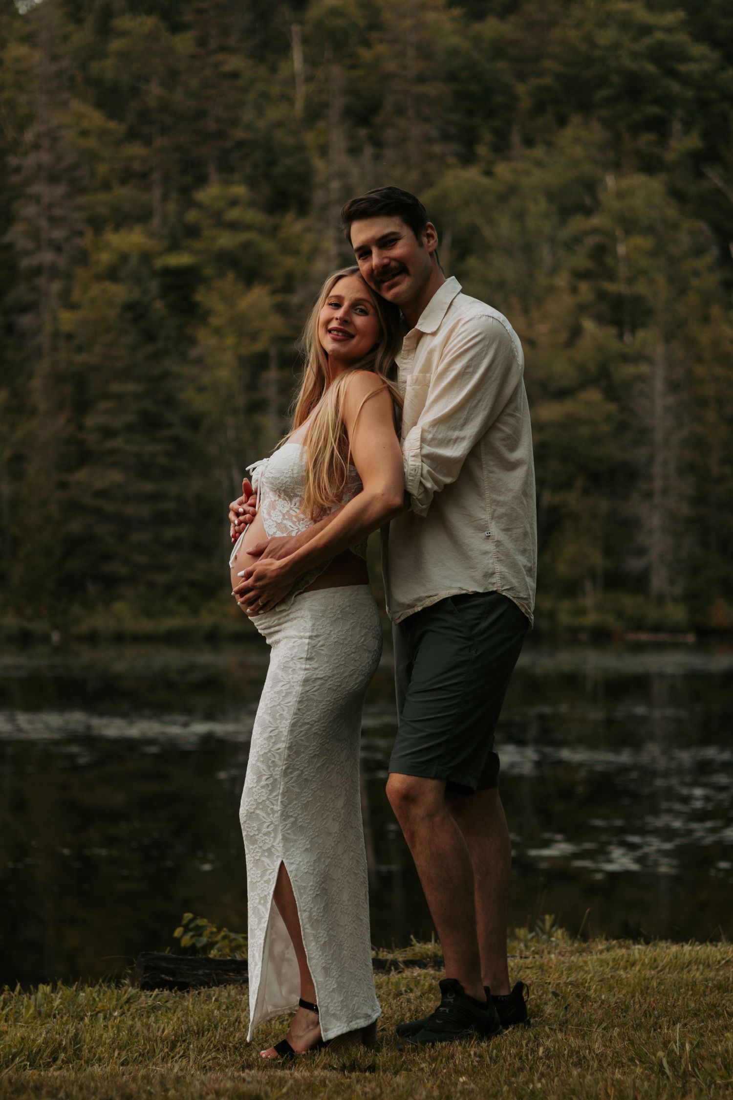 Couple shares intimate moment by lakeside during maternity photoshoot in white dress and casual attire.
