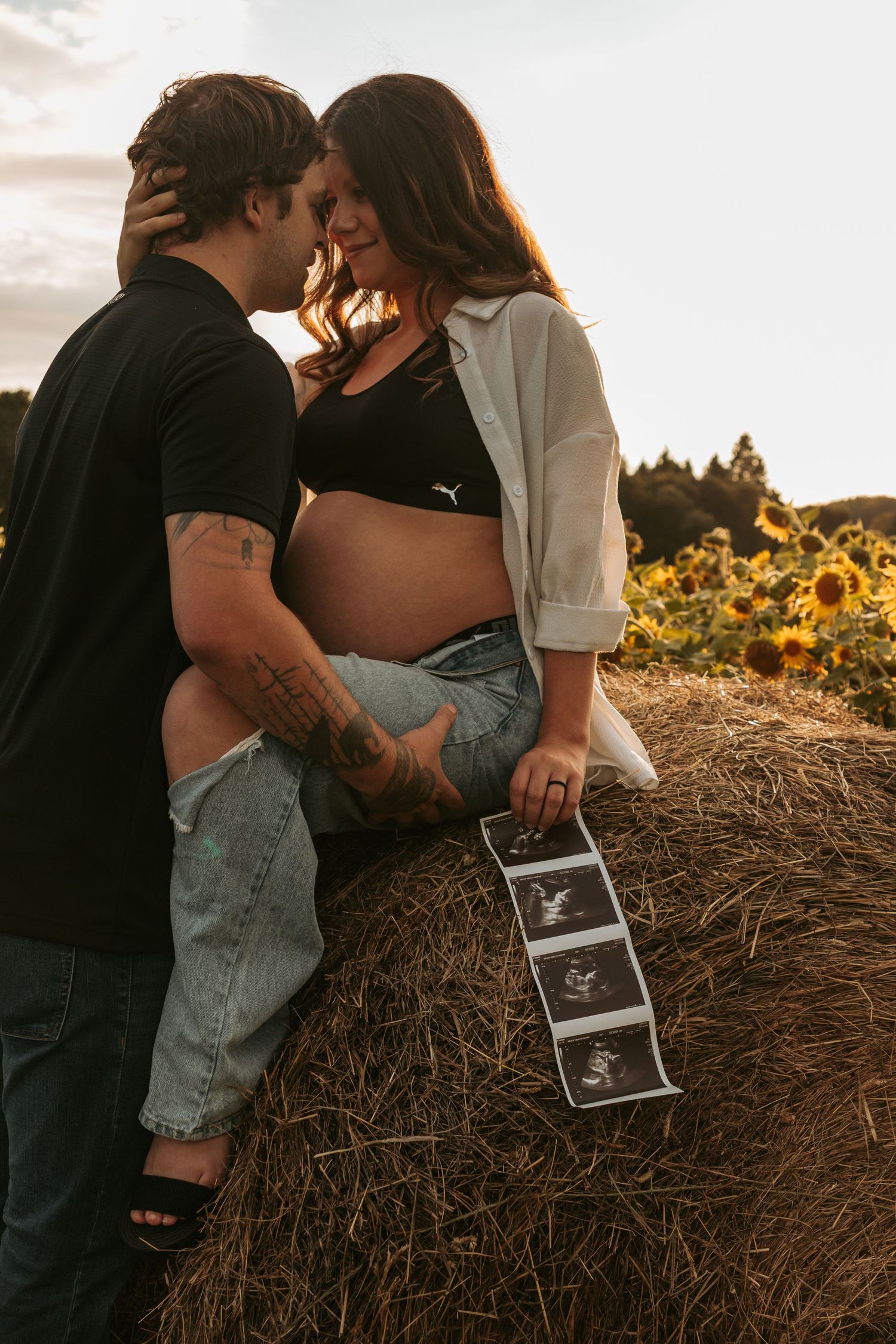 Couple poses with ultrasound photo against hay bale in sunflower field during sunset photography session.