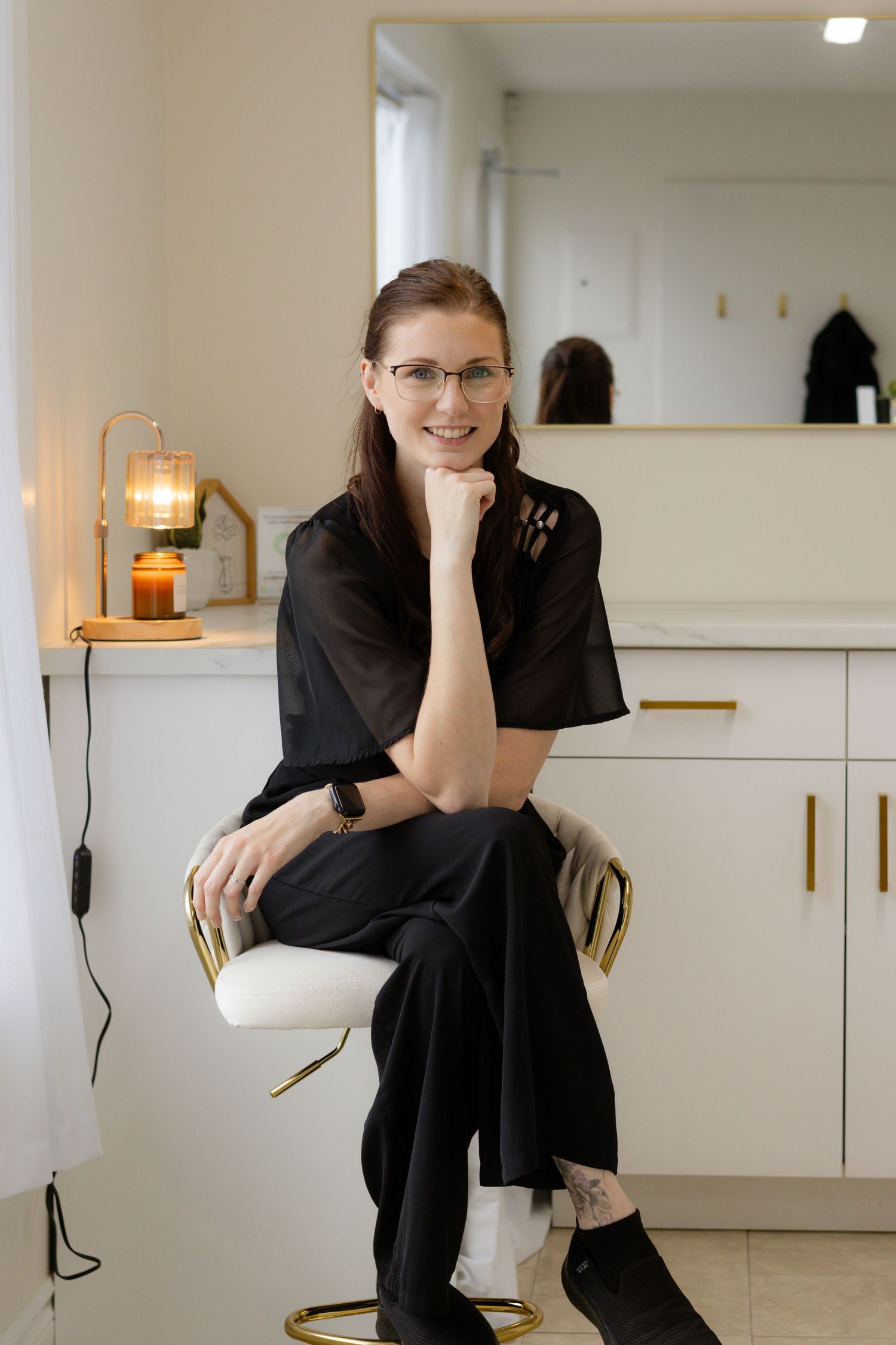 A smiling woman in black outfit and glasses sits on a gold stool in a modern salon interior.