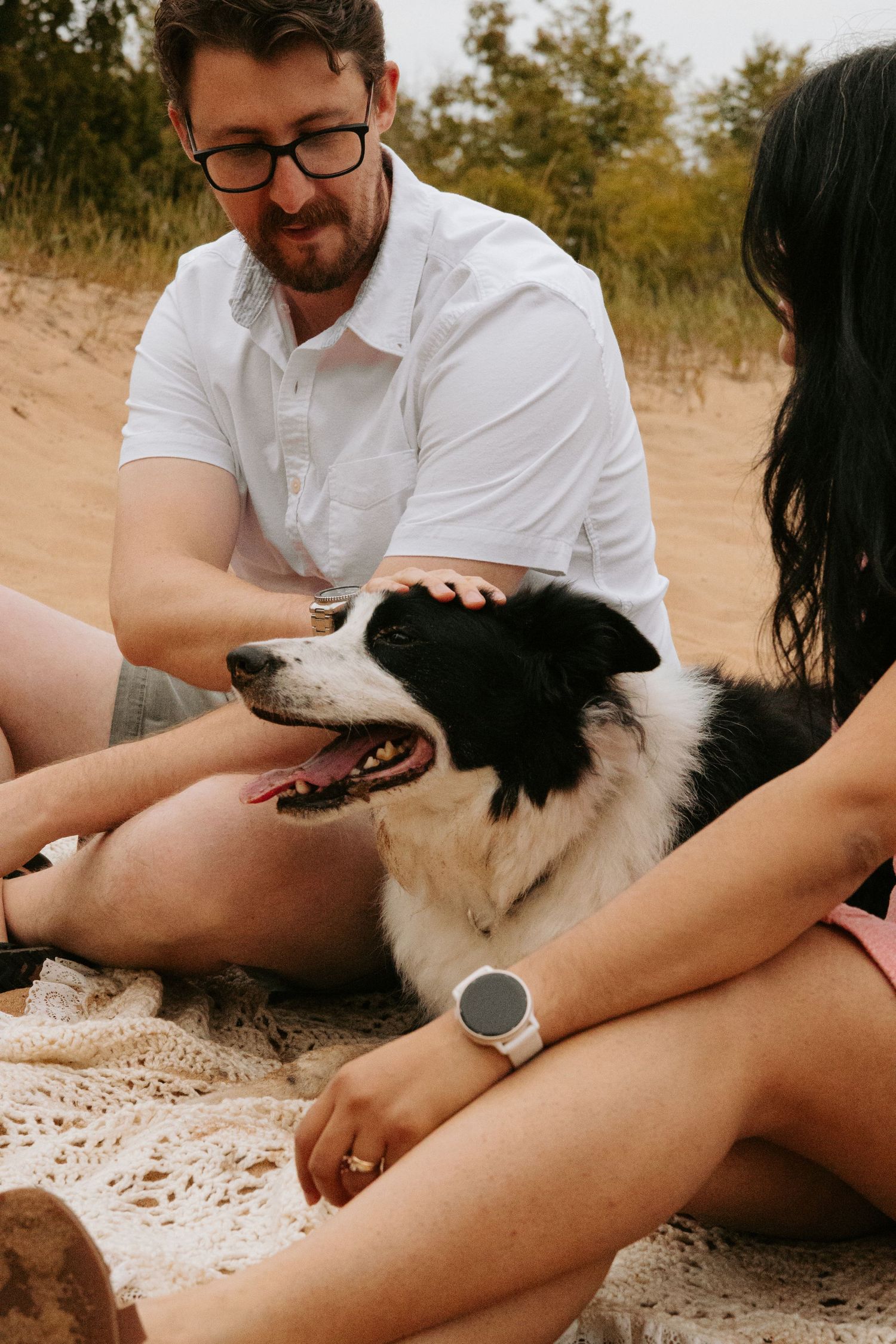 A couple pets their black and white dog while sitting on a blanket outdoors.