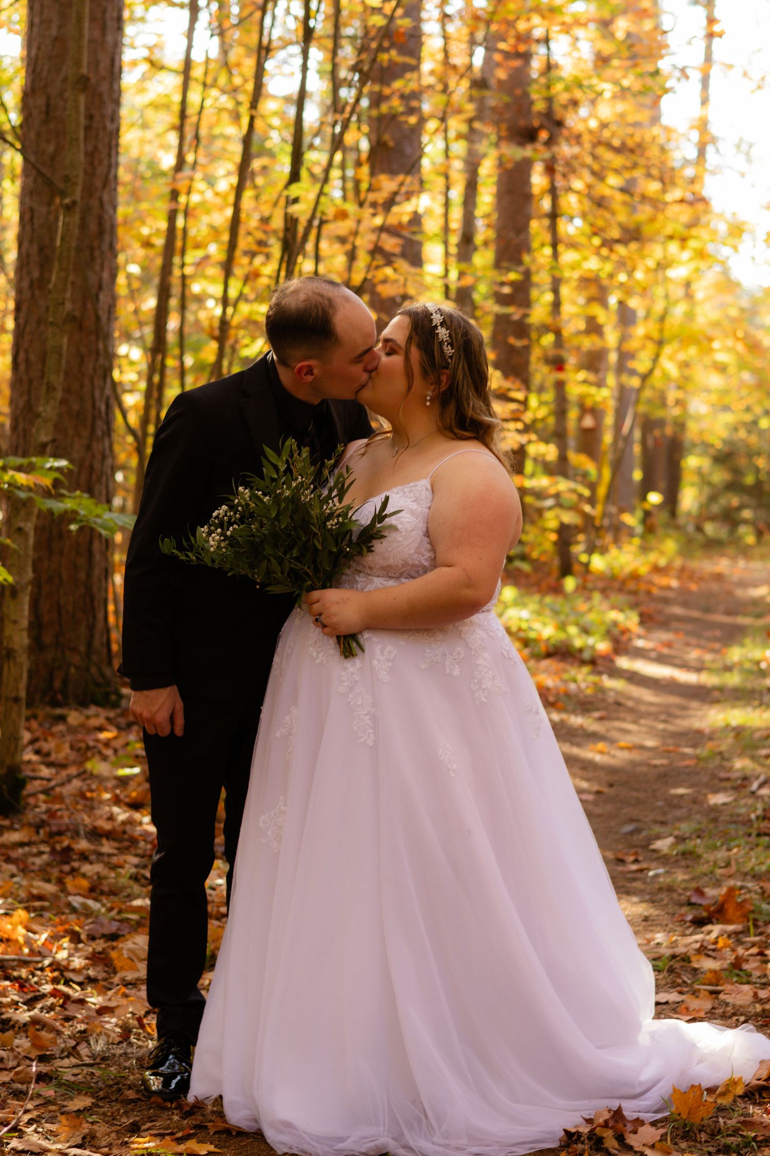 A romantic autumn wedding photo of a couple kissing on a forest path covered in golden leaves.
