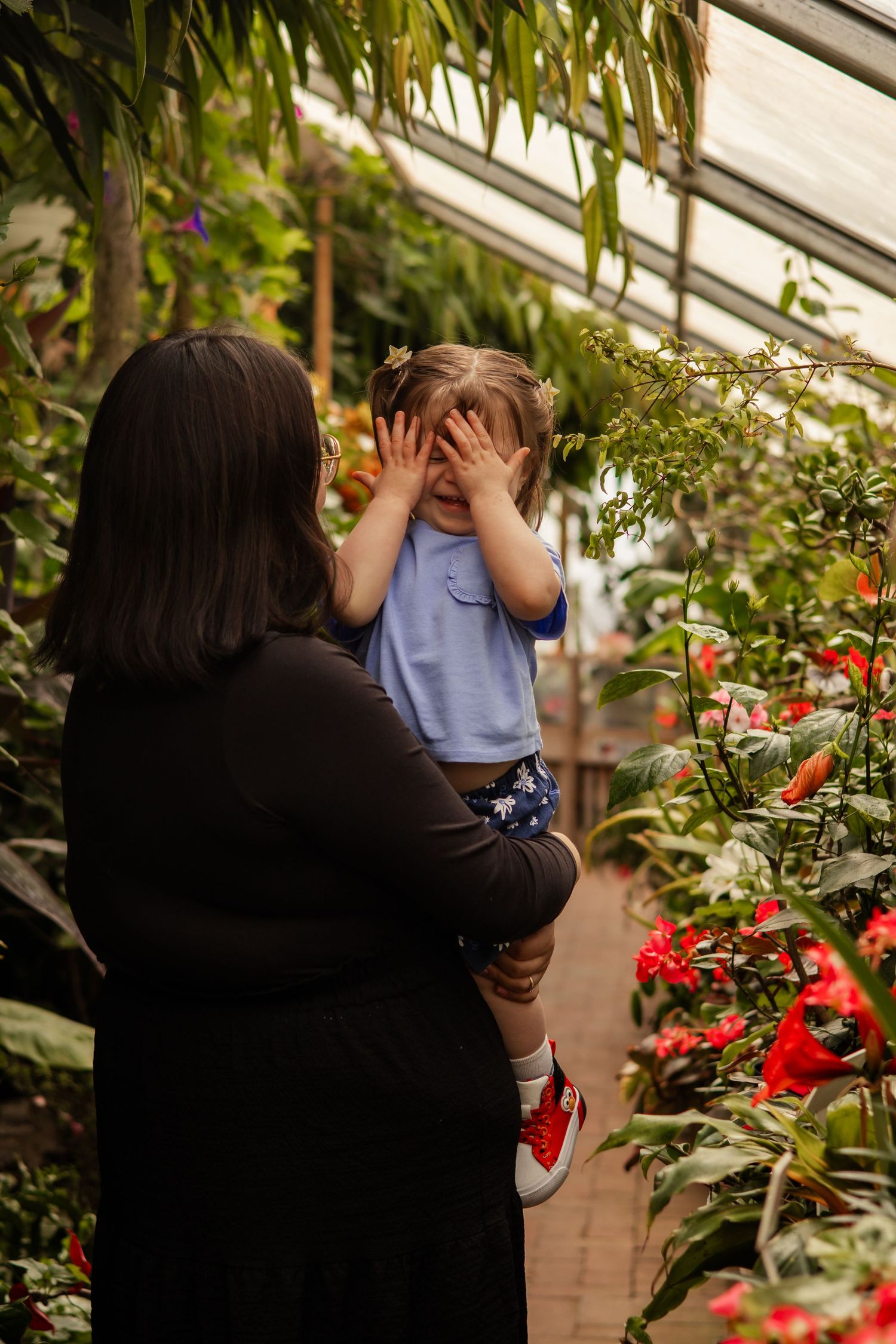 Mother carrying young child in a botanical greenhouse, toddler covering her face playfully.