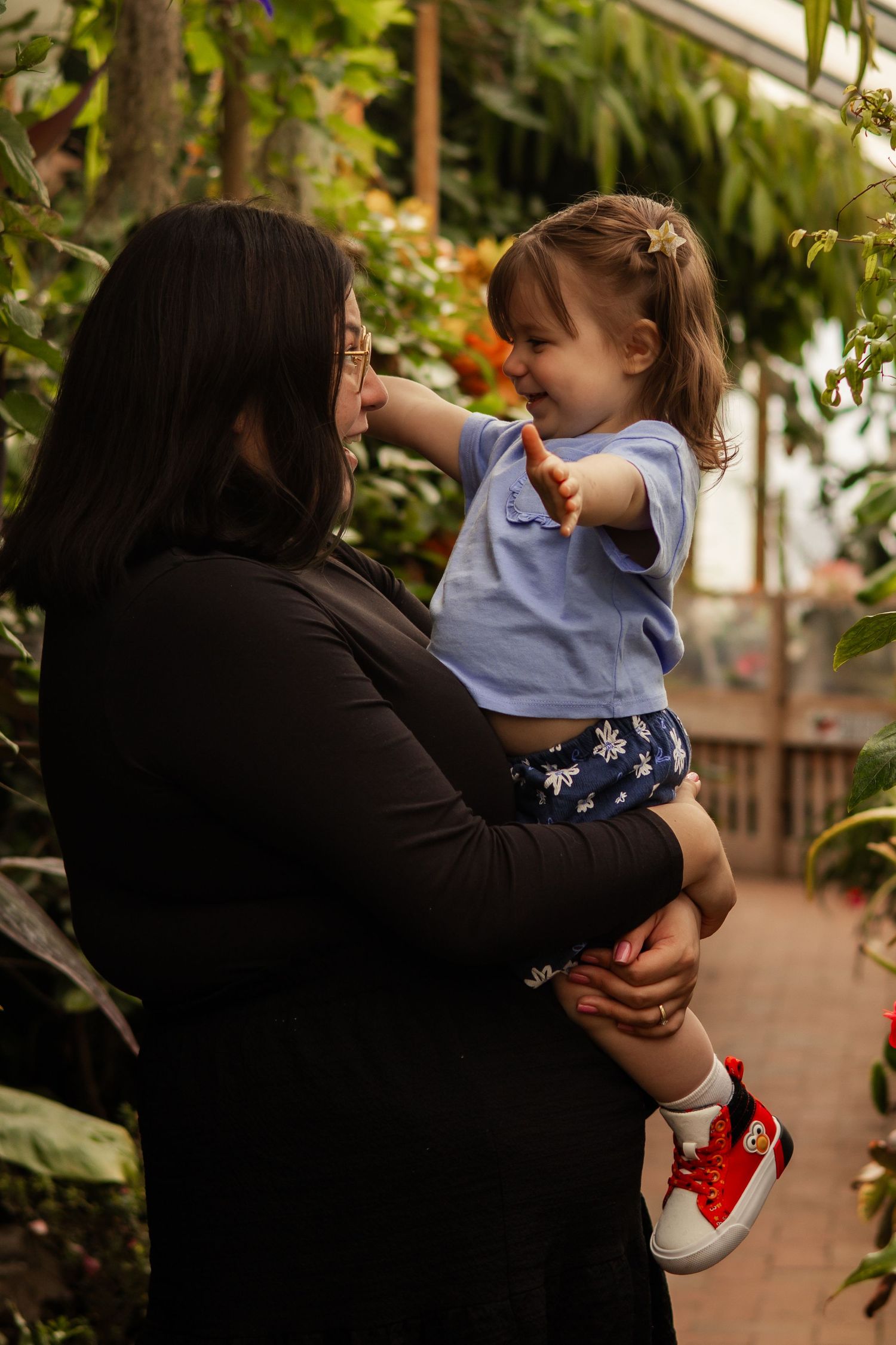 Woman holding a smiling toddler girl in a lush greenhouse, child pointing at colorful plants.