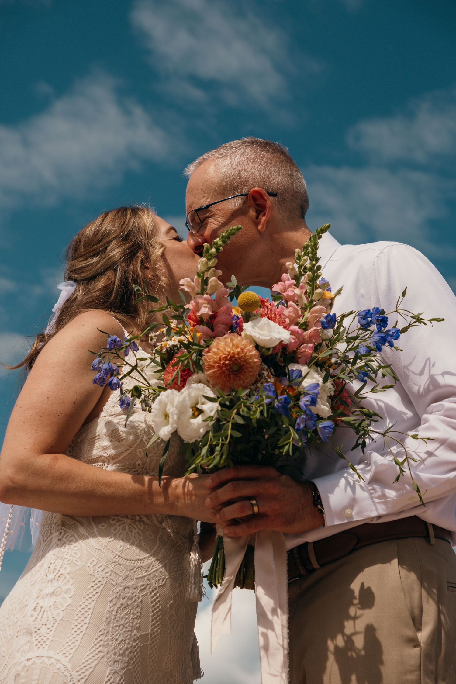 Romantic wedding couple embracing behind colorful floral bouquet against blue sky.