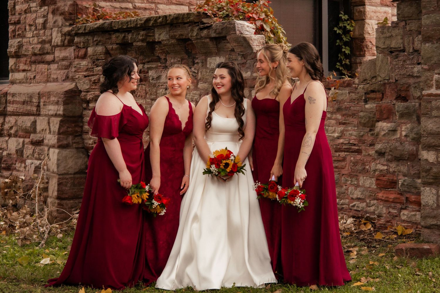 A bridal party in burgundy dresses poses together against a brick wall.