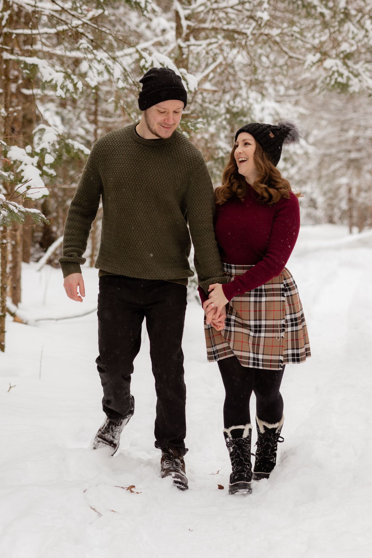 A couple walking together through snowy woods wearing winter clothing and plaid.