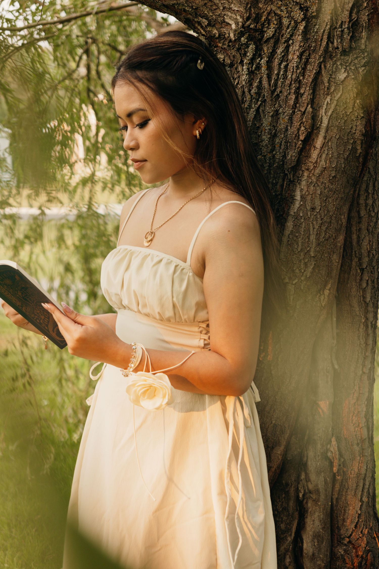 A dreamy portrait of someone in a white sundress reading against a tree trunk in soft natural lighting.