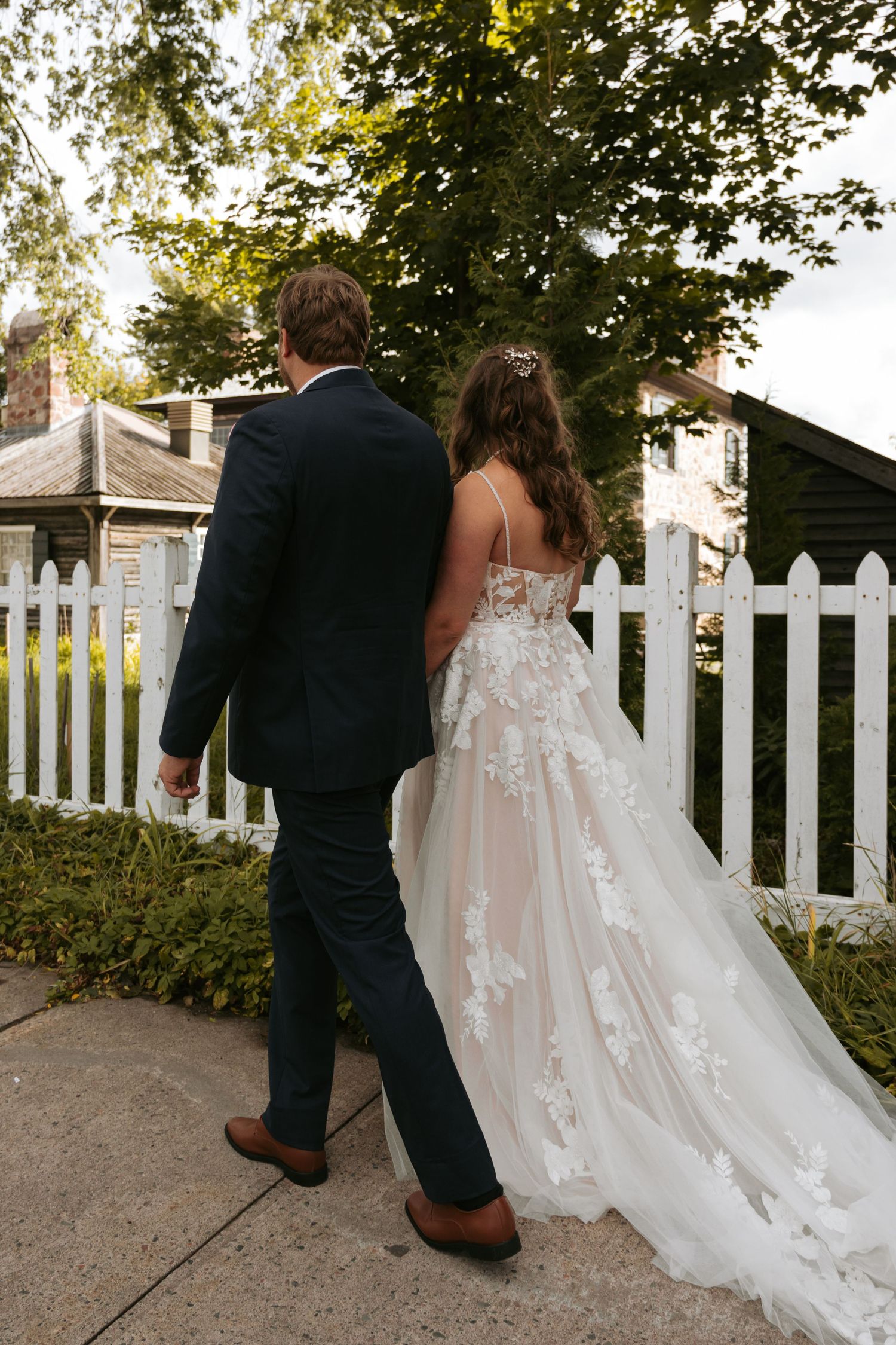 A couple walks along a white picket fence pathway during their wedding photoshoot.