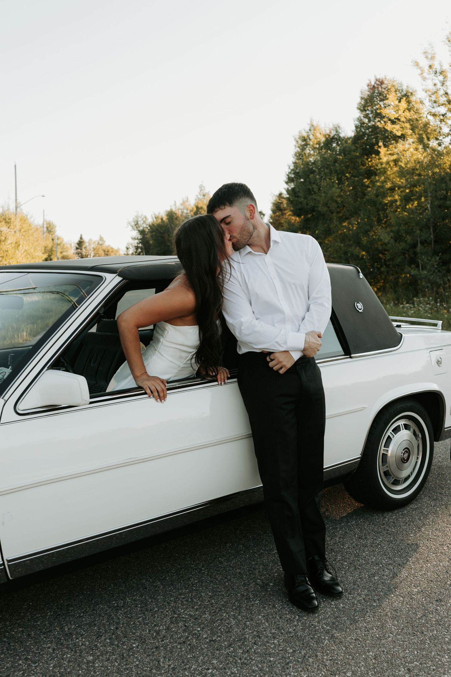 Stylish couple posing playfully against a classic white car at sunset.
