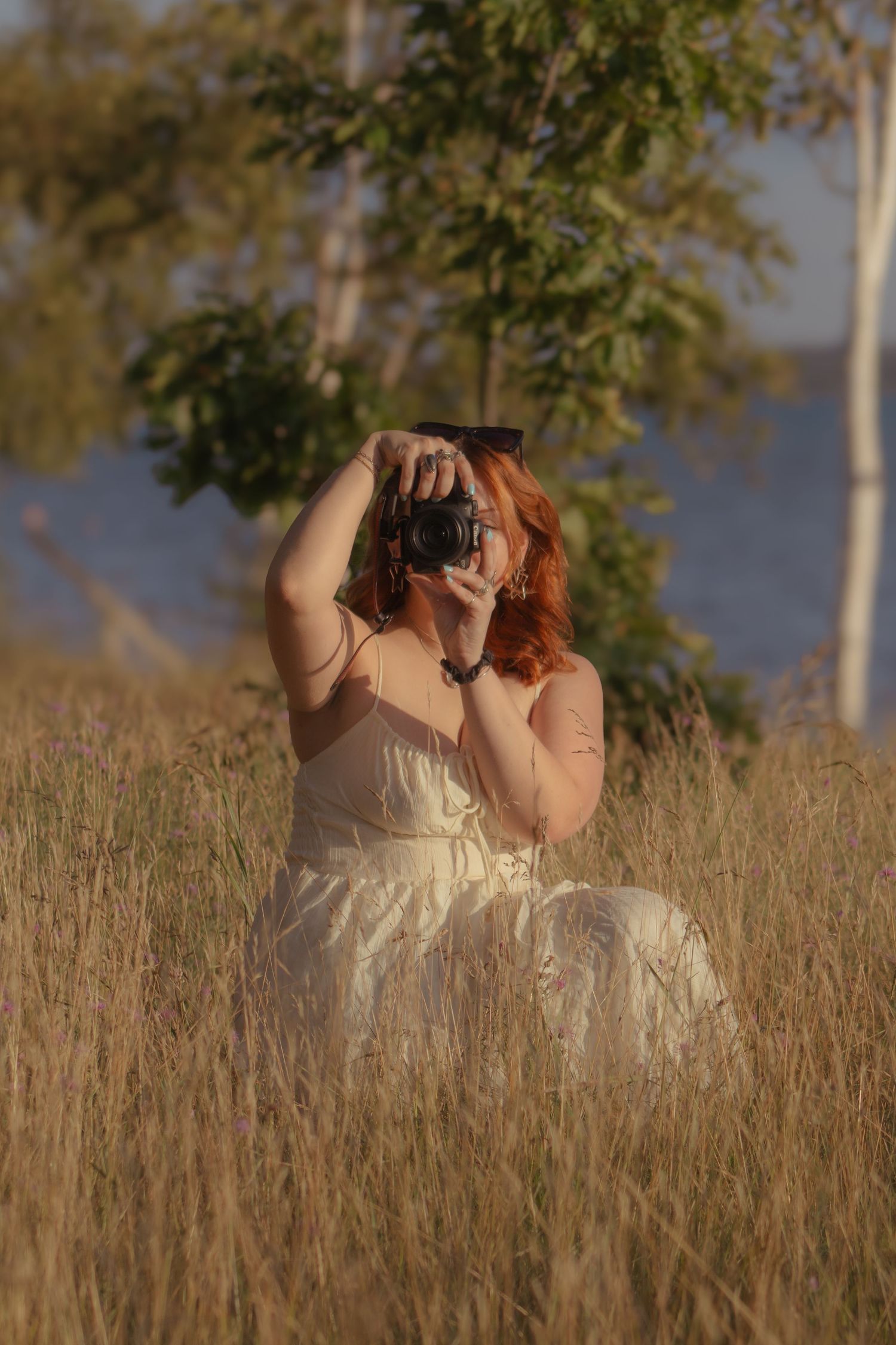 Red-haired woman in white dress sitting in tall grass, photographing with a DSLR camera outdoors.
