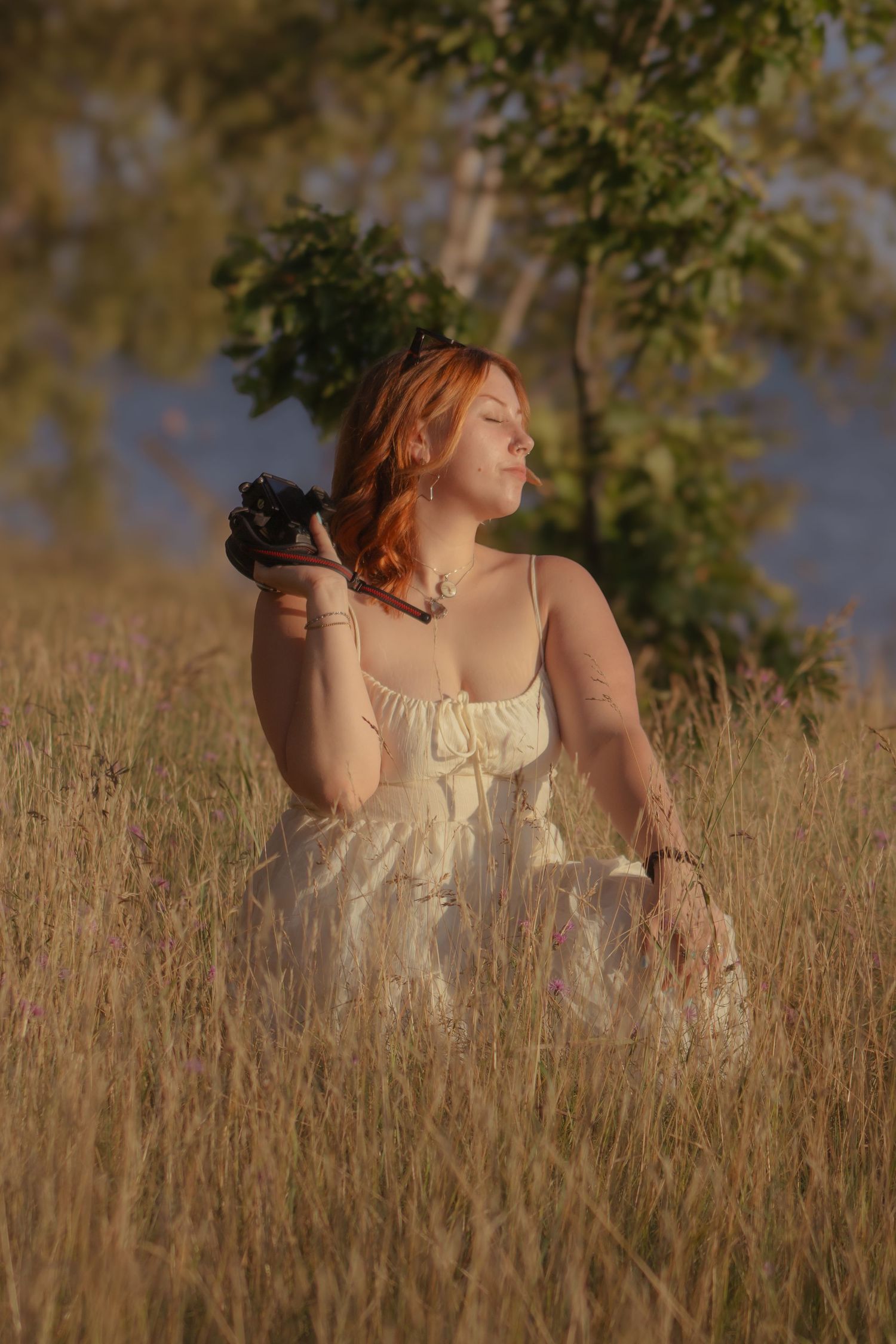 Red-haired woman in white dress holding camera sitting in golden meadow grass with trees in warm sunset light.