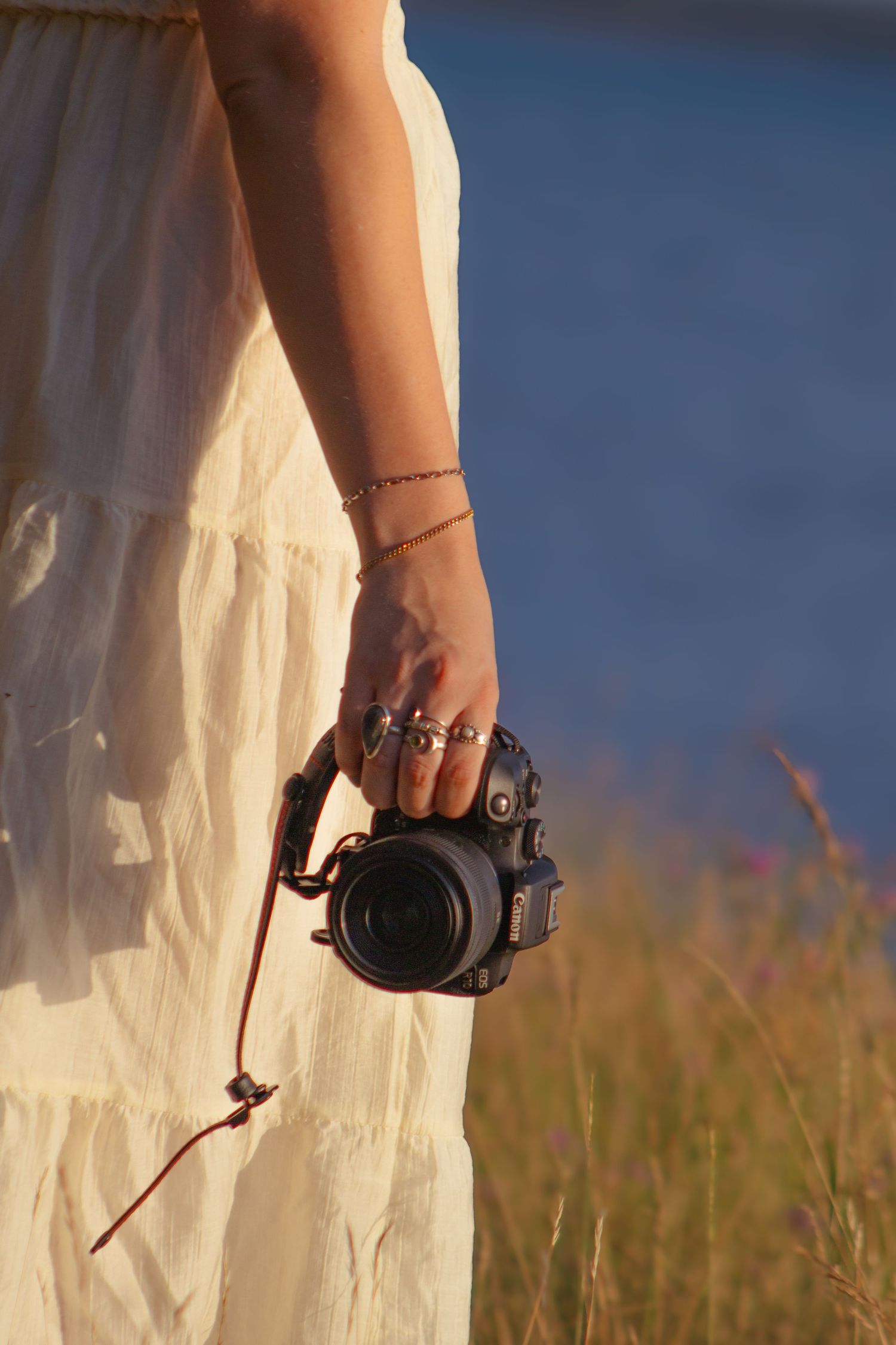 Woman in white dress holding a DSLR camera with rings and bracelet, standing near water in golden hour light.