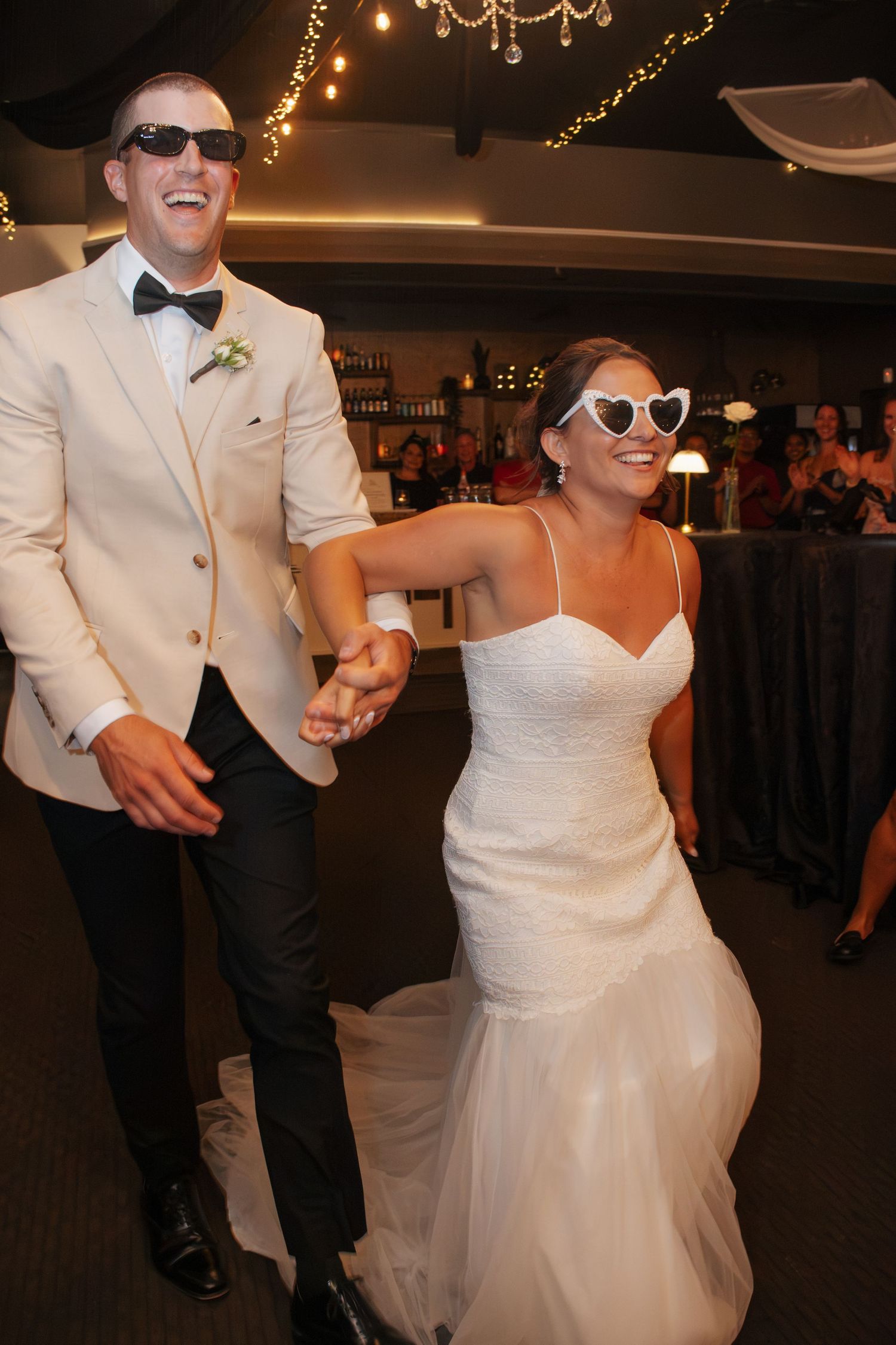 Bride and groom wearing sunglasses laughing and holding hands during wedding reception entrance.