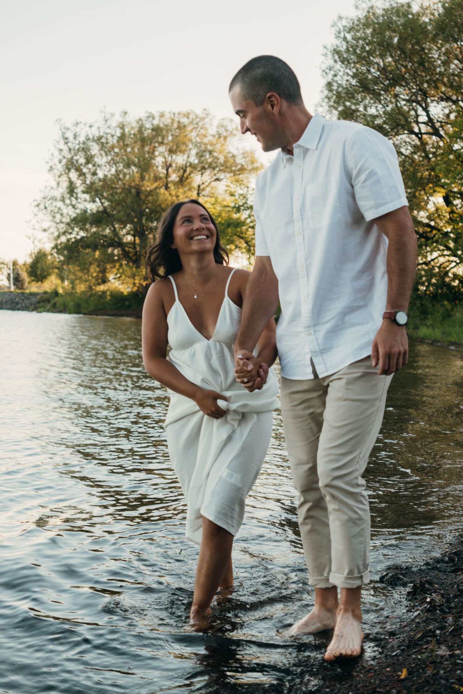 Couple in white outfits playing in shallow water during sunset photoshoot.