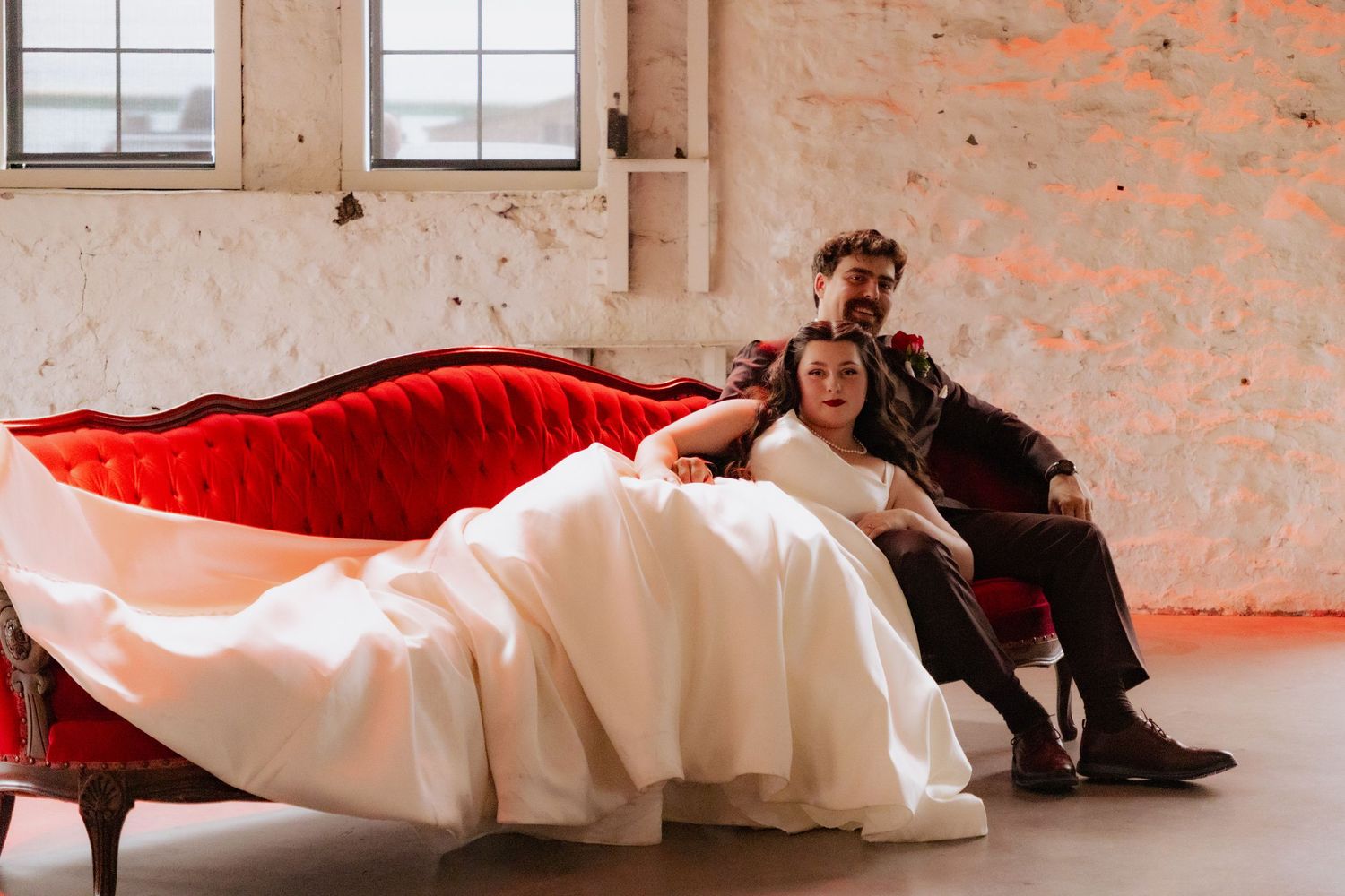 A couple relaxes on a vintage red chaise lounge in an industrial-style venue.