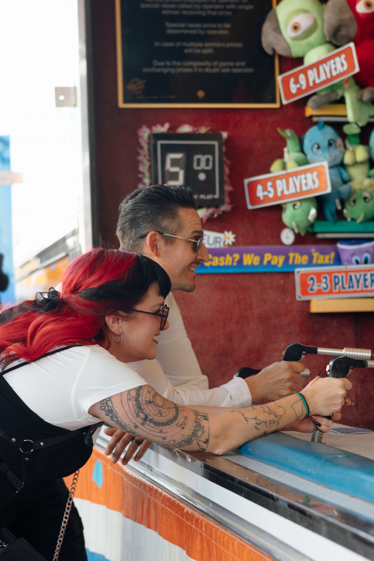 Couple with tattoos playing a carnival game at an amusement arcade, woman with red hair leaning on counter.