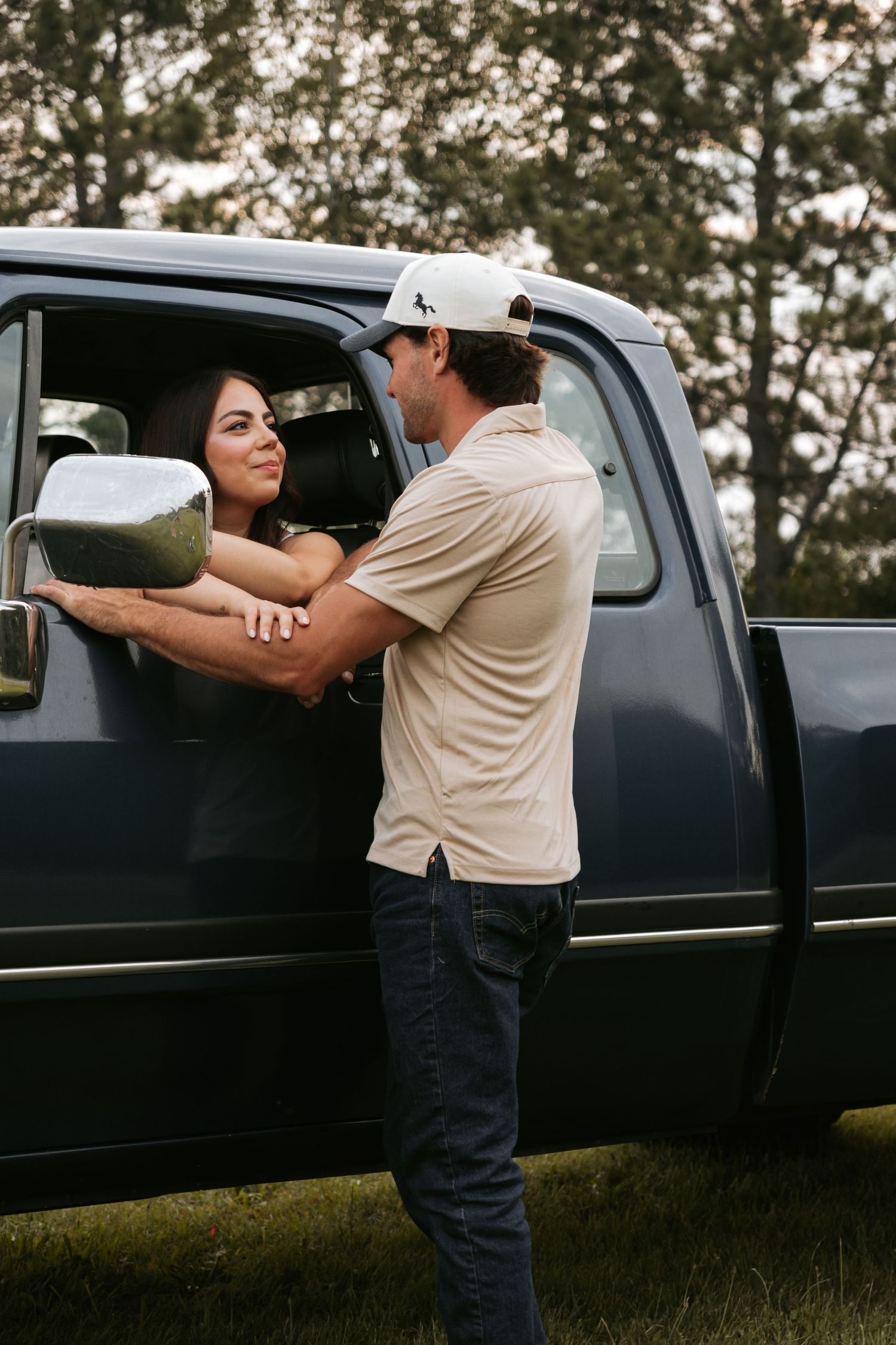 Two people lean against a black pickup truck in a field at dusk.