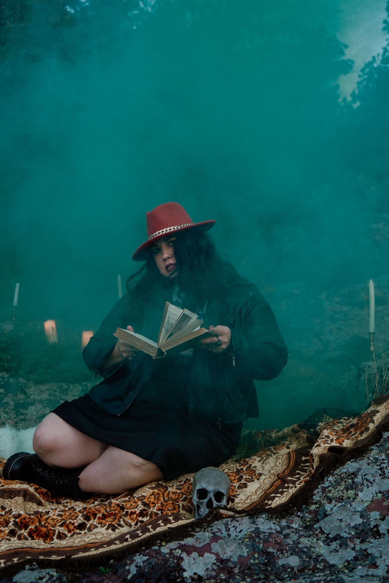Dark moody photoshoot featuring person in hat reading on blanket with decorative skull.