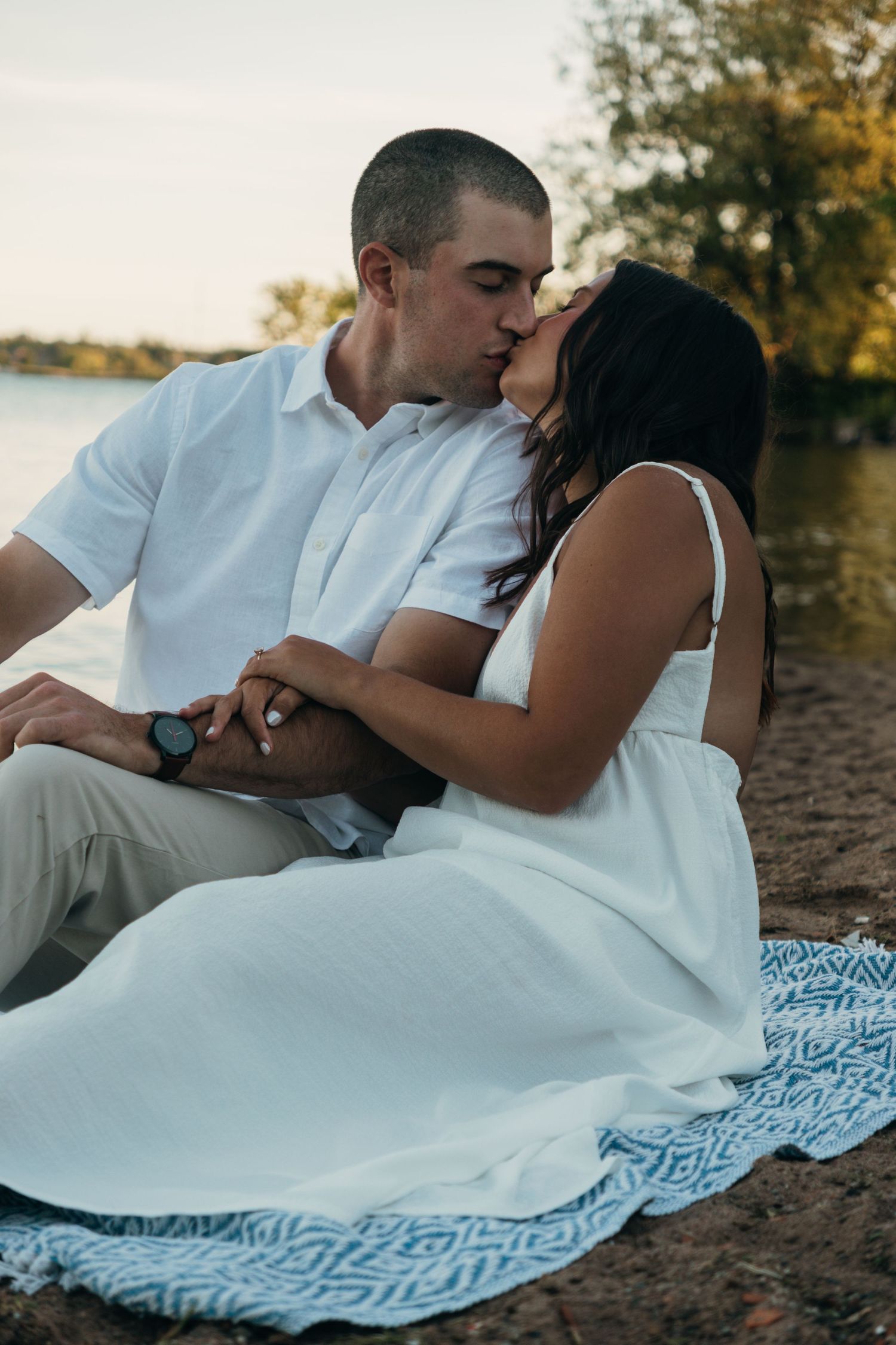 Couple in white attire sharing intimate moment on patterned blanket by waterfront.