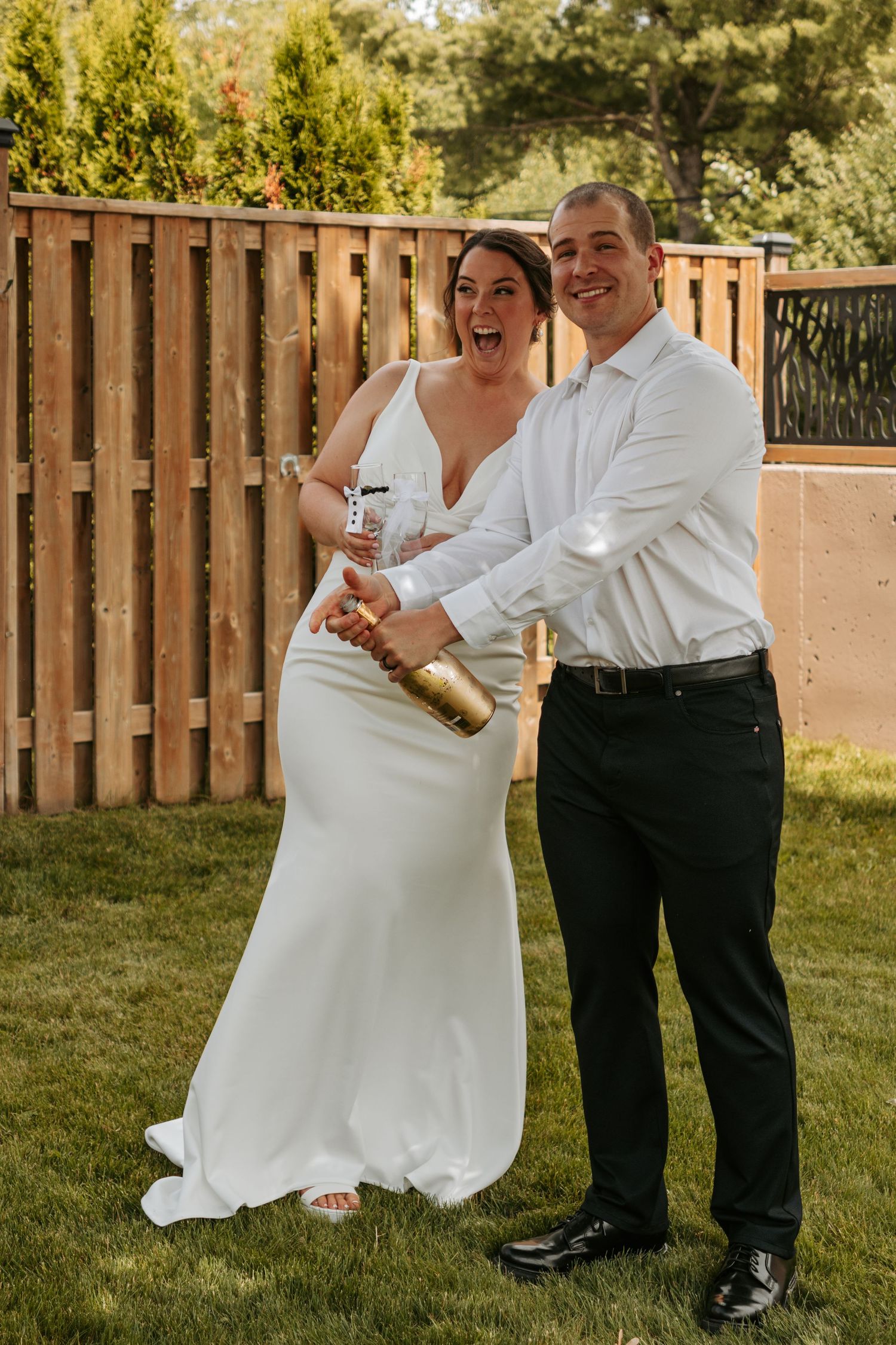 A couple celebrates their wedding day with champagne while standing on a grassy lawn.